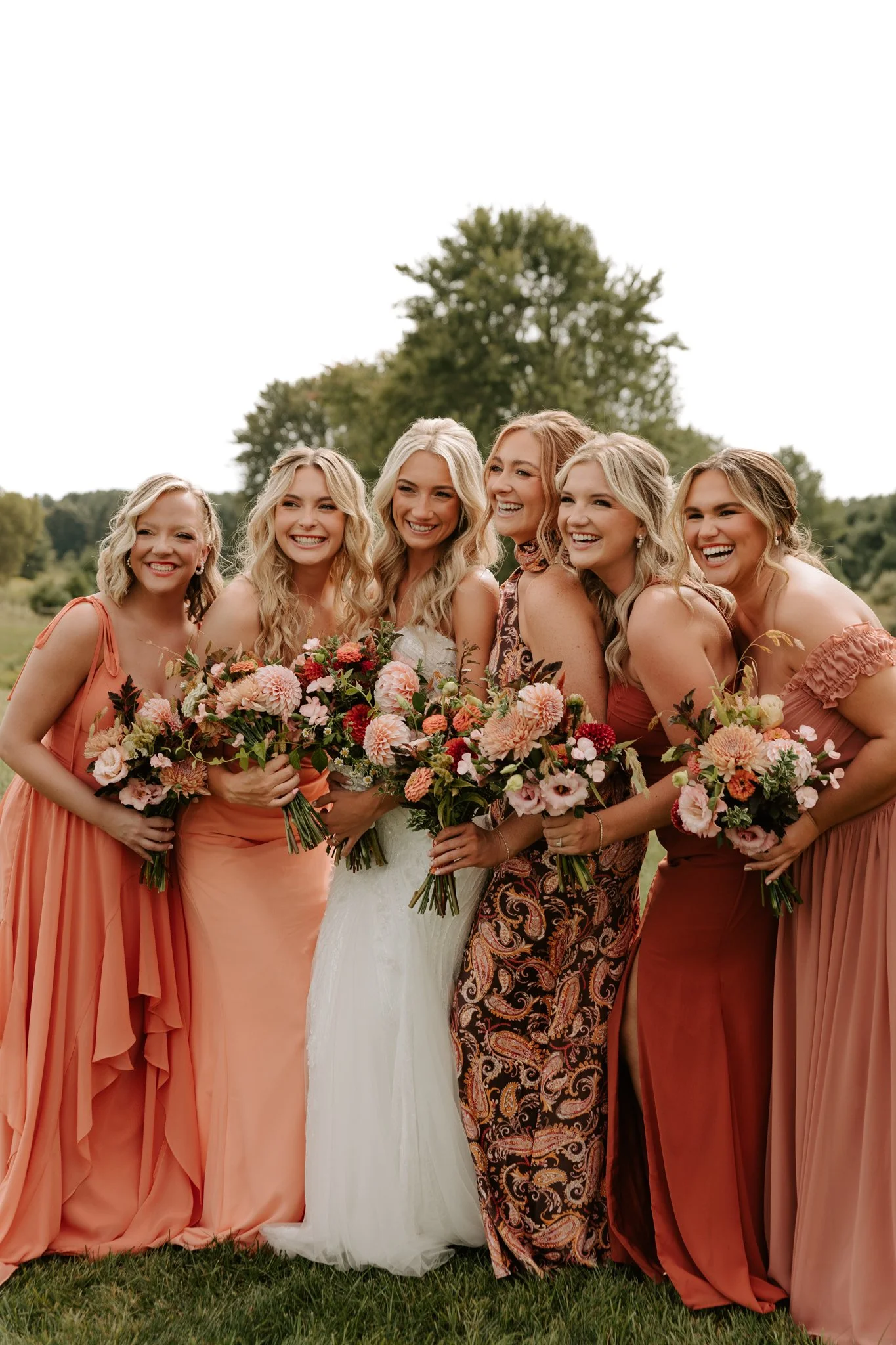 Group of six women in colorful dresses holding bouquets of pink and red flowers, smiling outdoors on a grassy field with trees in the background.