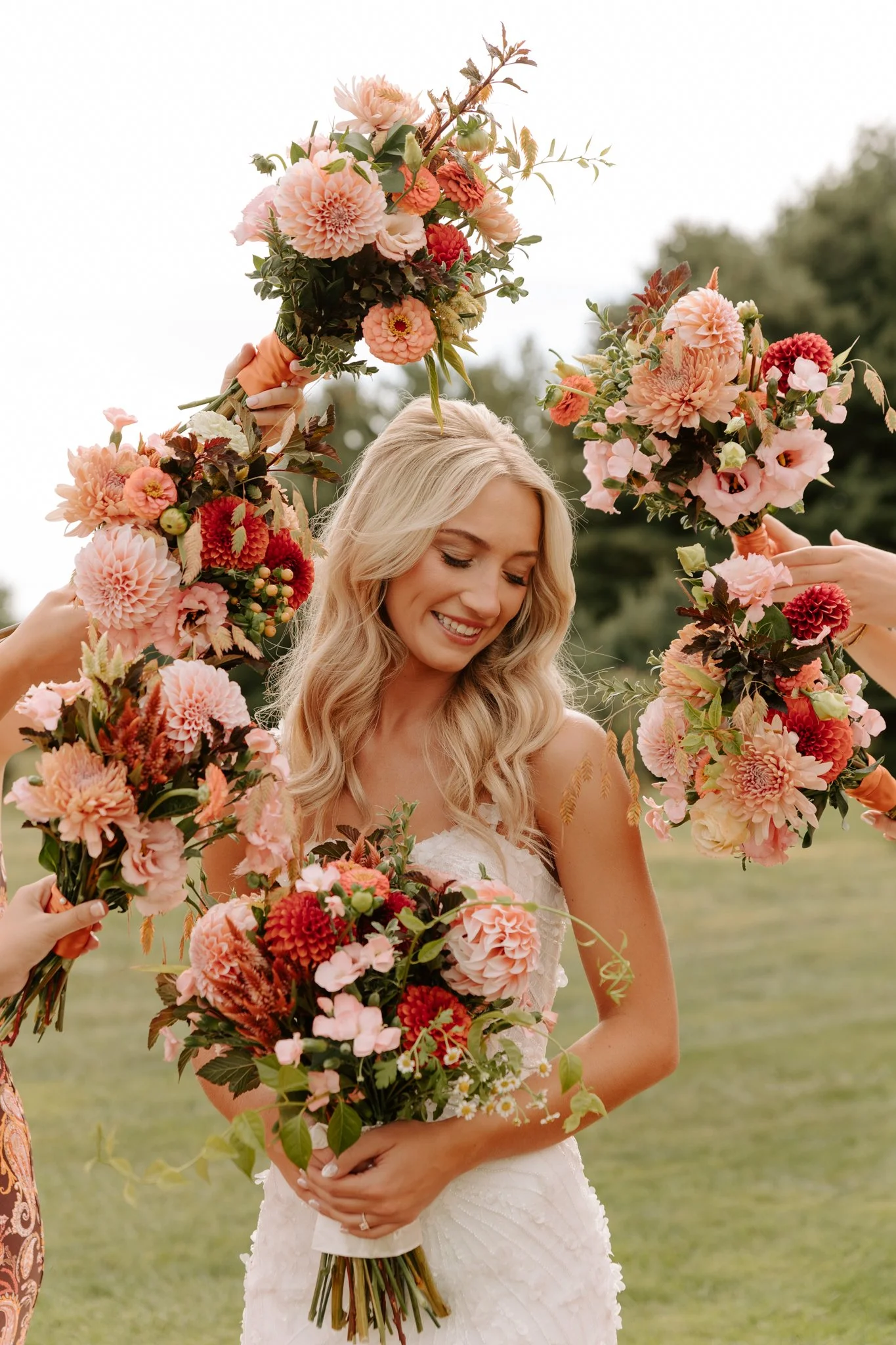 A woman in a white wedding dress holding a bouquet of pink and red flowers, smiling, as she is surrounded by others holding matching flower arrangements.