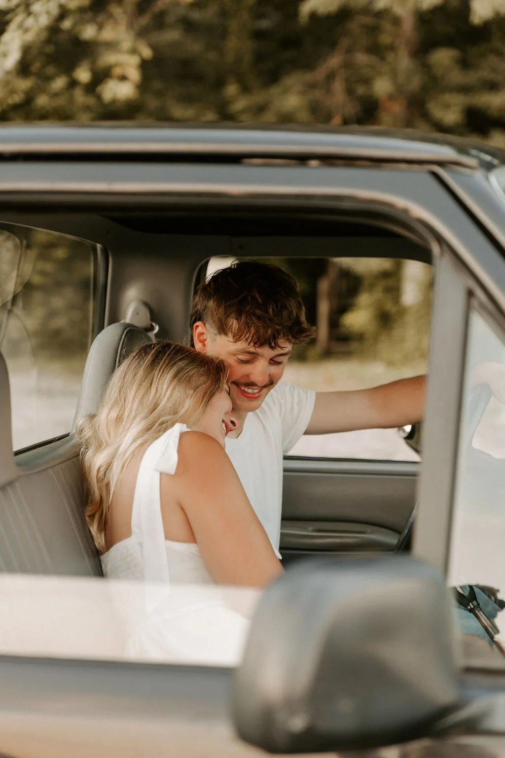 A smiling young man and woman laughing together inside a vintage vehicle, with the woman resting her head on the man's shoulder, surrounded by a nature-filled background.