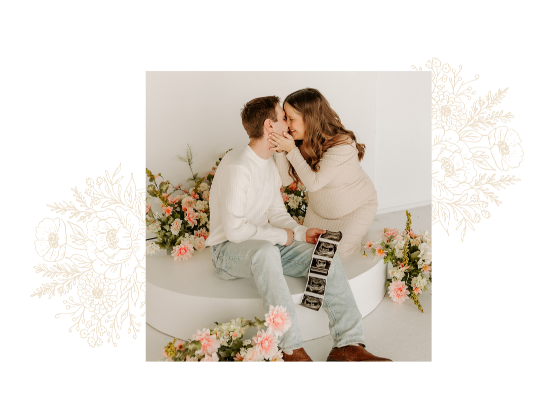 A couple is sitting on a white curved bench with a backdrop of pink and white flowers. The woman, who appears to be pregnant, leans in to kiss the man, who is holding ultrasound images. Both are dressed in light-colored clothing, and the scene has a soft, romantic atmosphere.