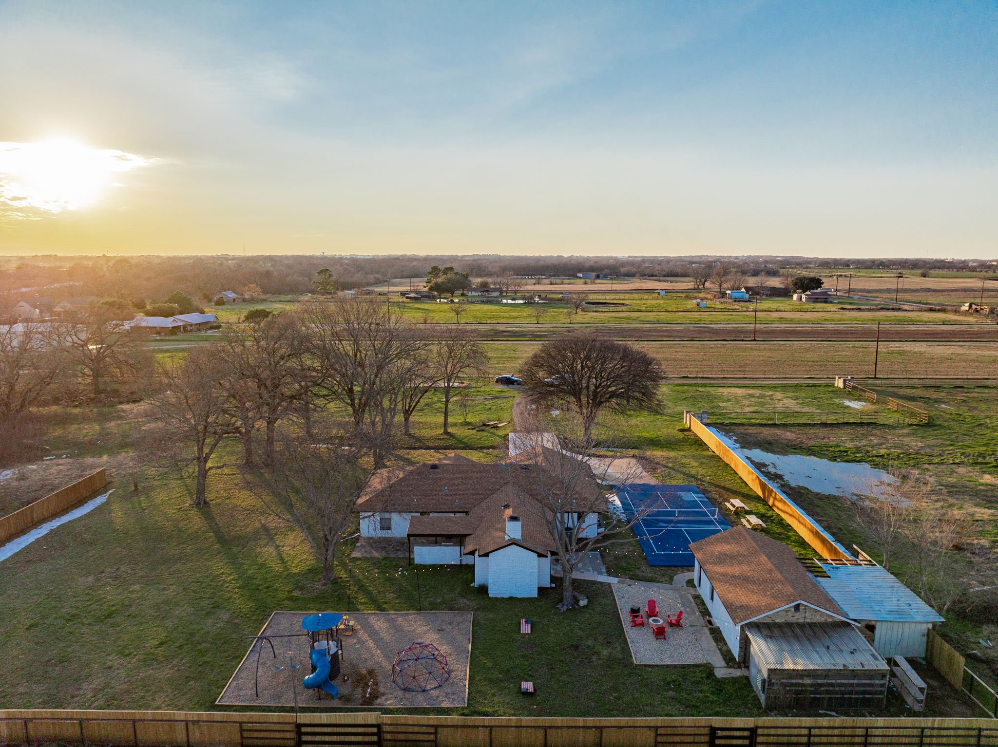 Aerial view of a rural backyard with trees, a house, a swimming pool with a cover, a children’s play area with a slide and climbing dome, and an outdoor seating area with red chairs. The yard is fenced and surrounded by open farmland, with a few scattered buildings and trees. The sun is setting on the horizon.