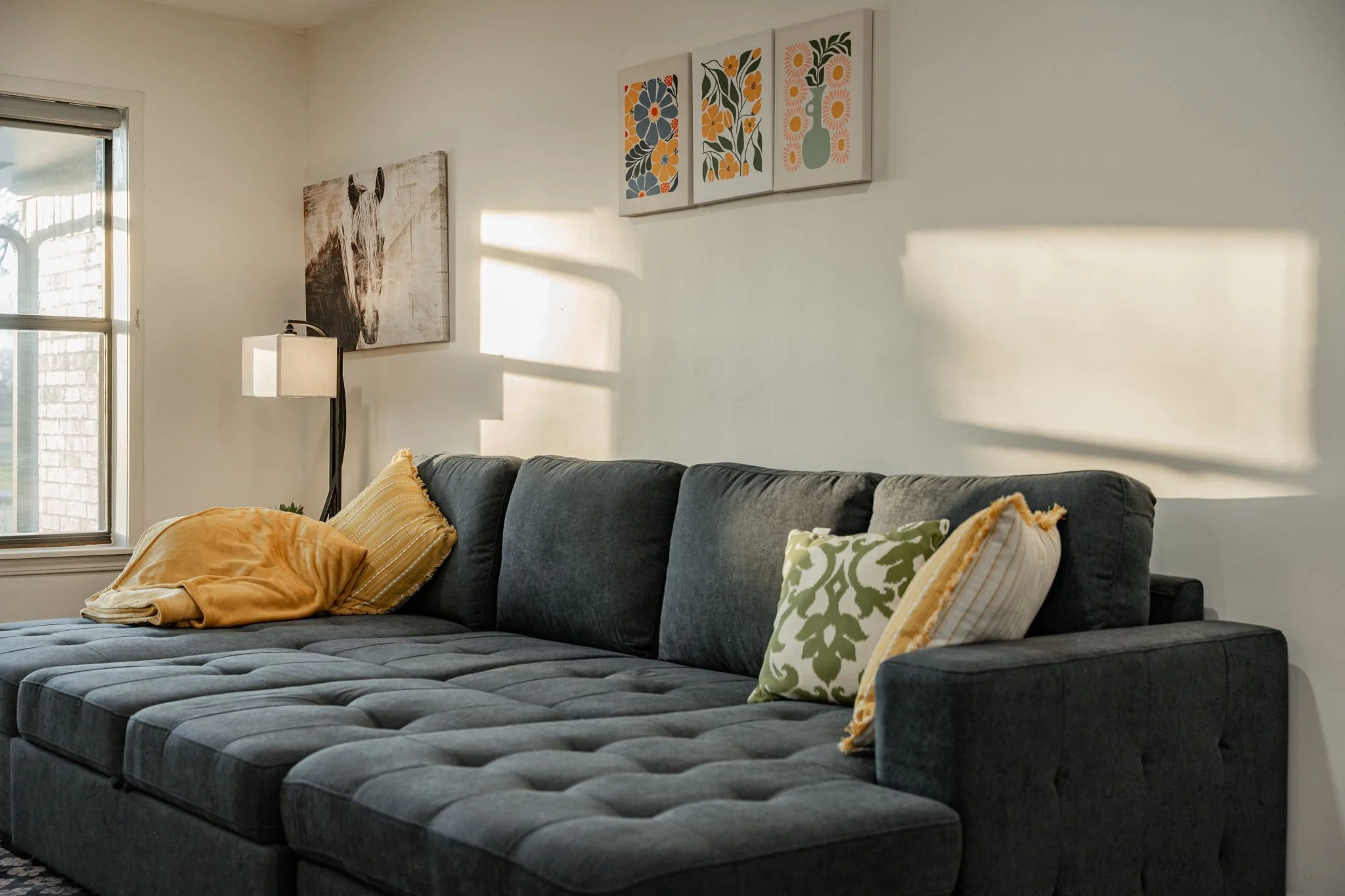 A living room with a gray sectional sofa, decorative pillows, wall art, a standing lamp, and sunlight casting shadows on the wall.