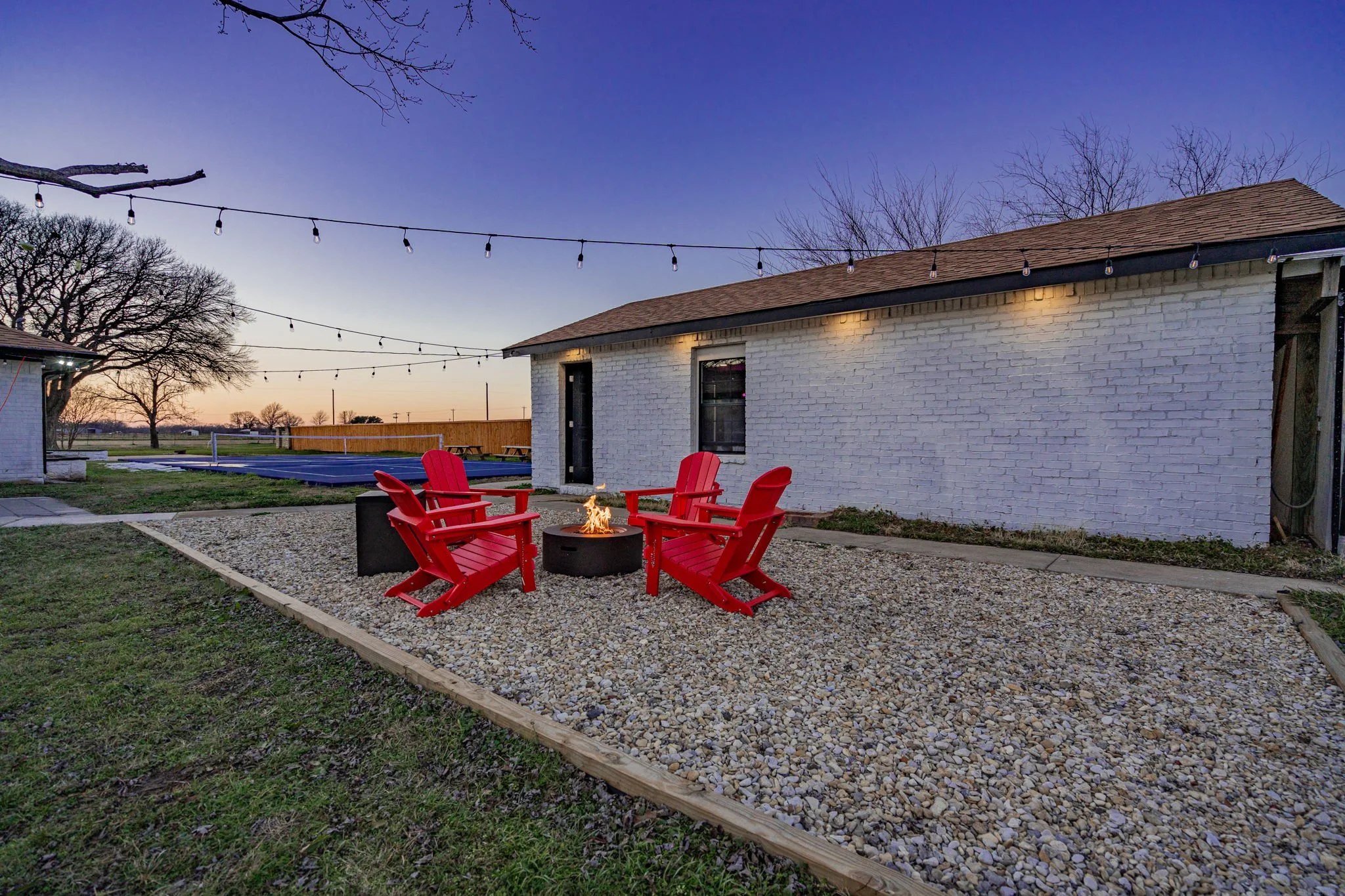 Backyard patio with four red Adirondack chairs around a fire pit on a gravel surface, string lights overhead, a white brick building, a fenced area with a swimming pool, and bare trees at dusk.