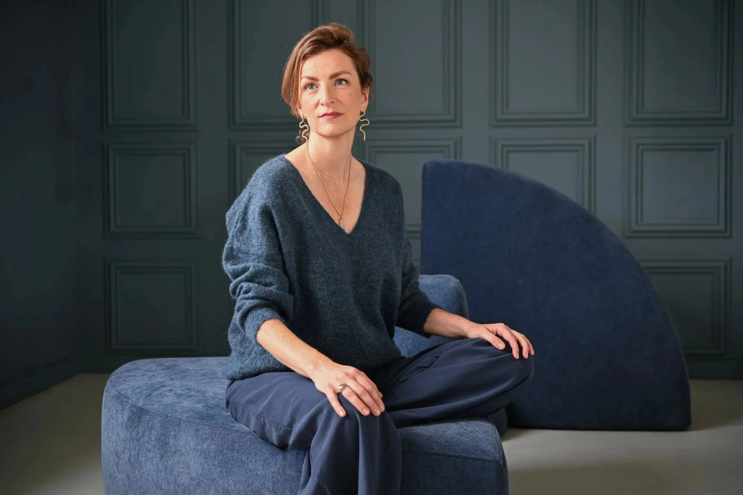 A woman with red hair sitting cross-legged on a dark blue cushioned seat in front of a dark paneled wall, wearing a navy sweater and dark pants, with jewelry including earrings and a necklace.