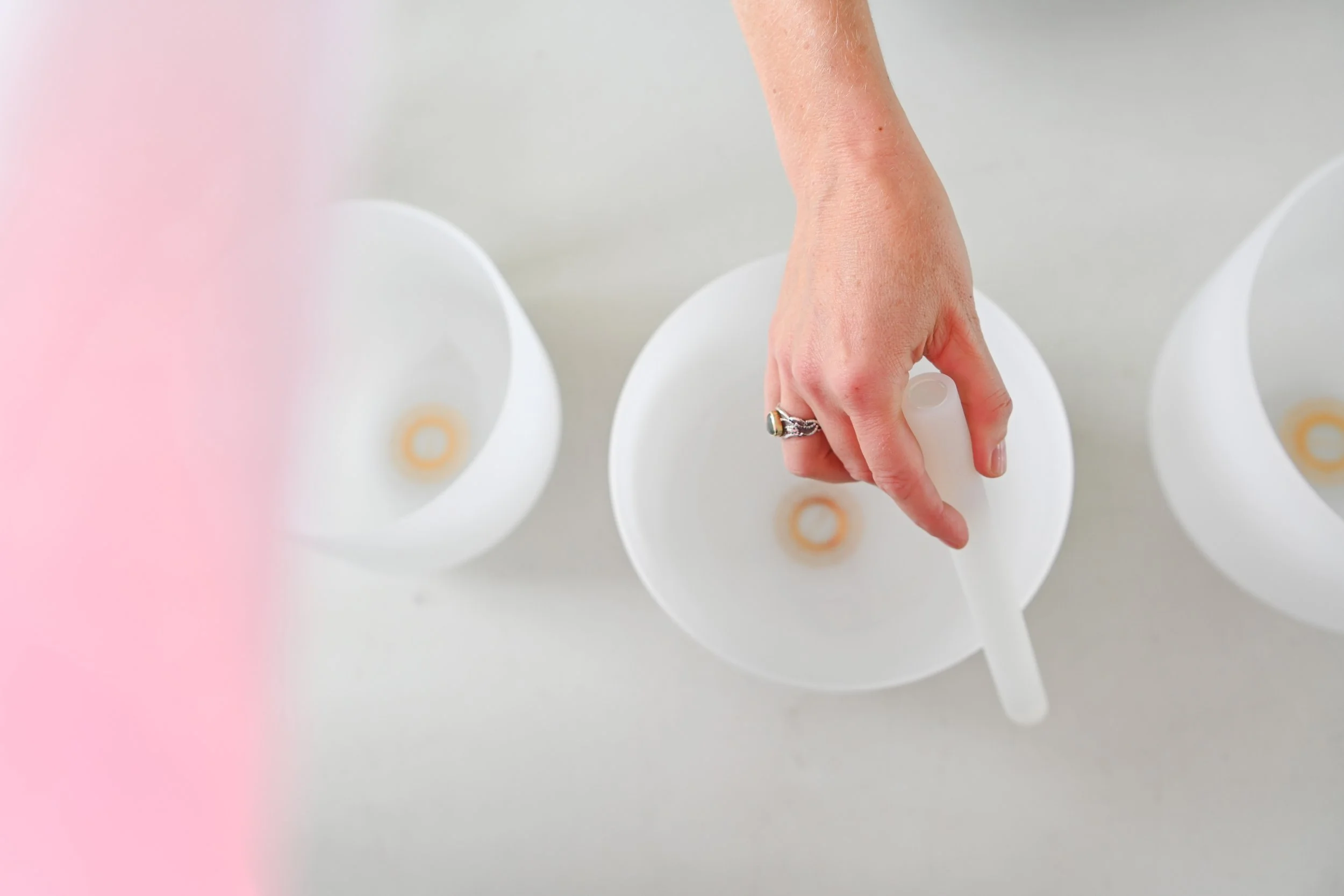 A hand with a silver ring picking up a white spoon from a white bowl on a light-colored surface, with two other bowls nearby.