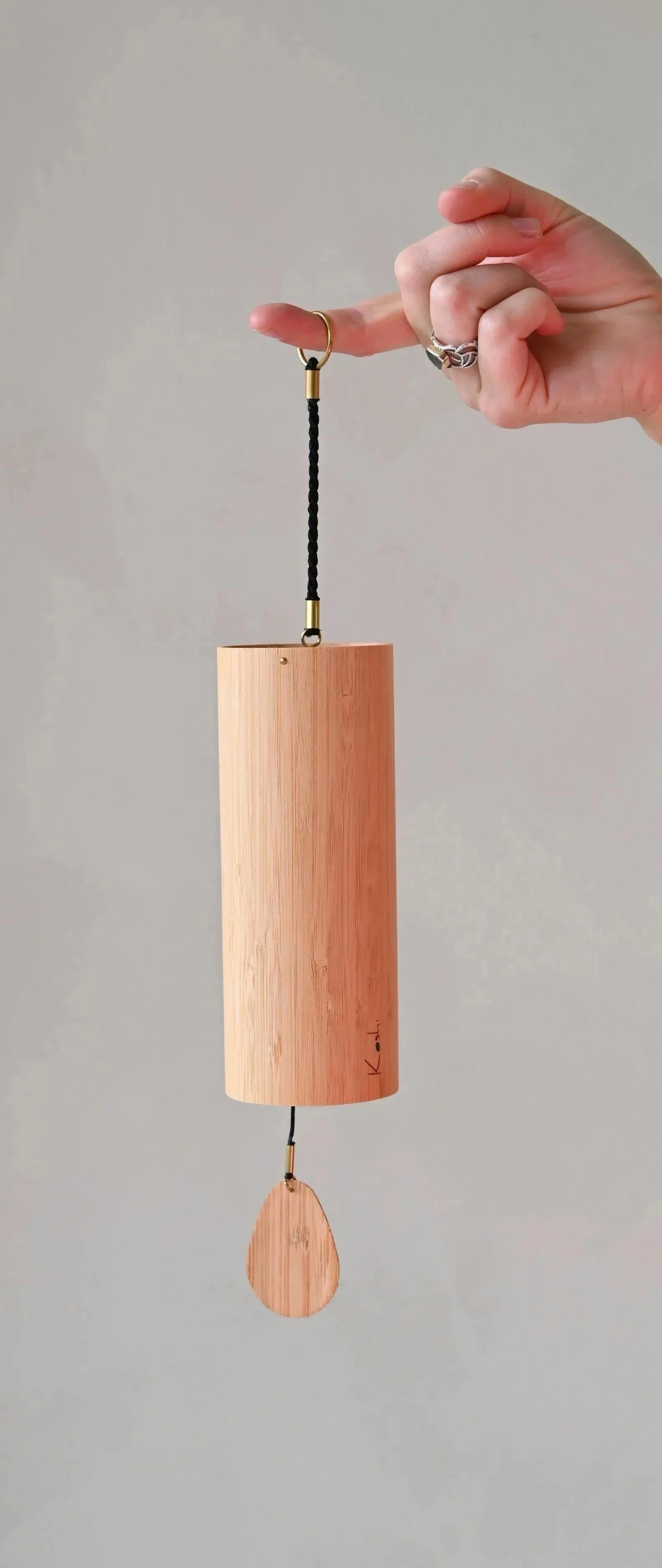 A person's hand holding a wooden wind chime with a black cord hanging from a gold ring, with a plain gray background.