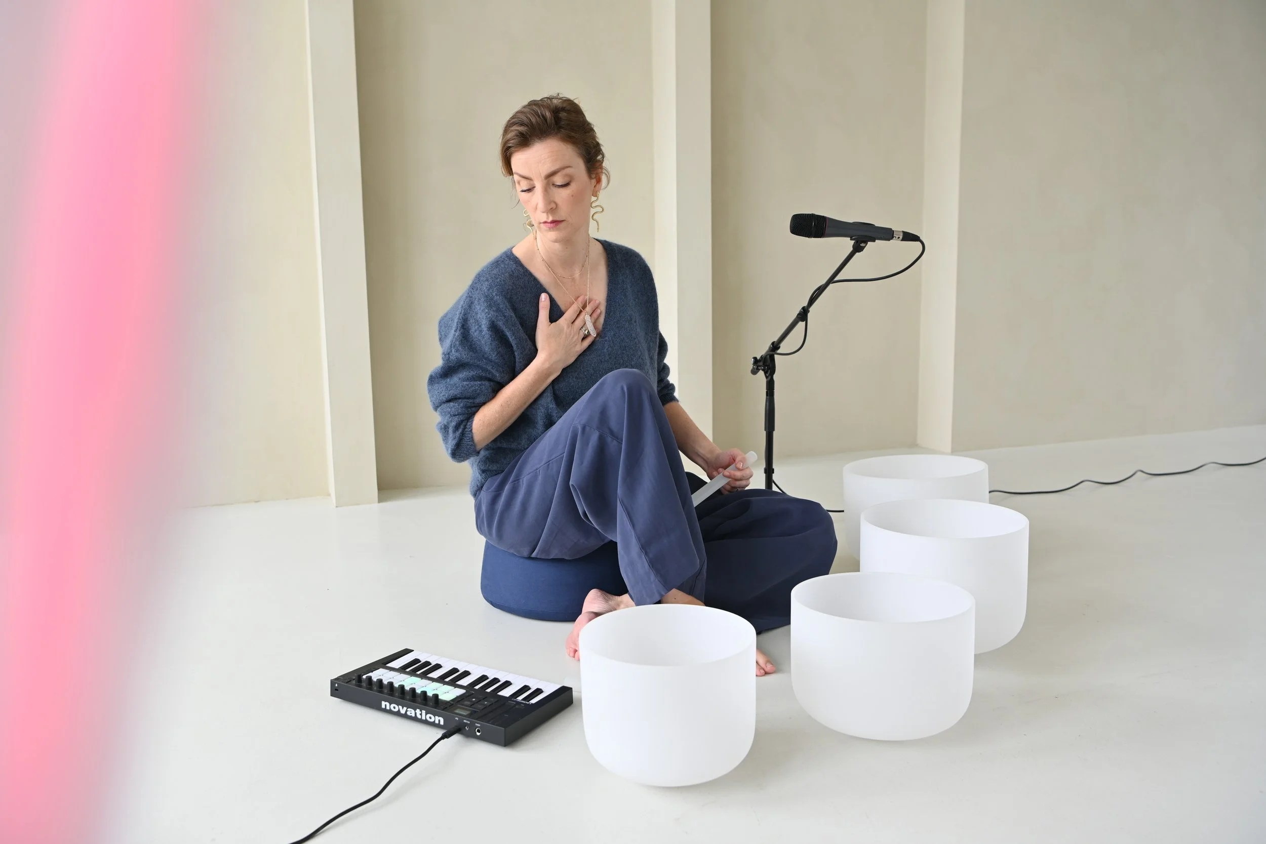A woman sitting on a blue cushion, surrounded by white singing bowls, with a microphone and a small keyboard nearby, in a minimalistic room.
