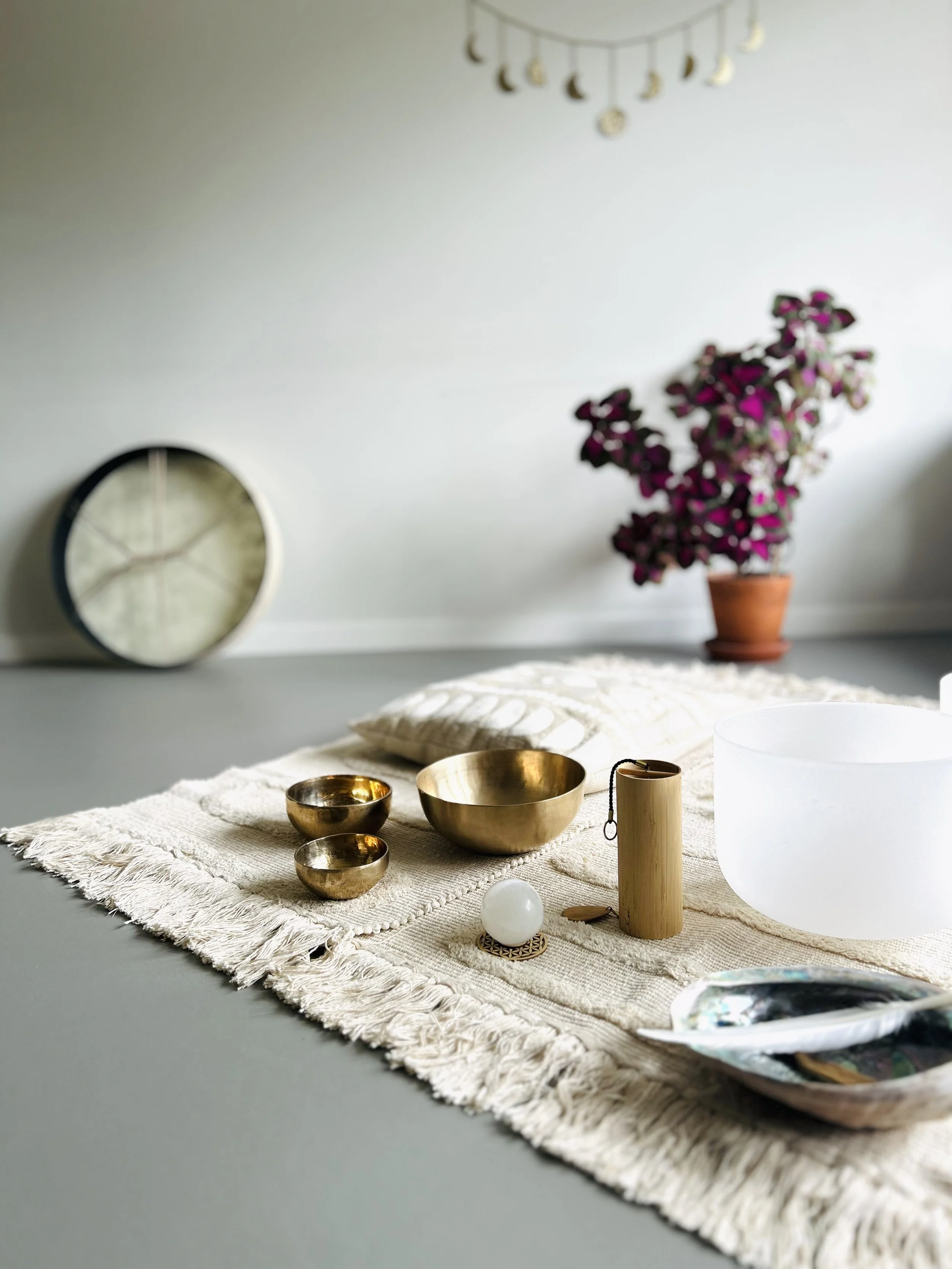 Bronze singing bowls, a white sphere, a wooden wind chime, a white chrystal singing bowl, and big seashell, on a fringed cloth, with a potted purple flowering plant and an ocean drum leaning against a wall..