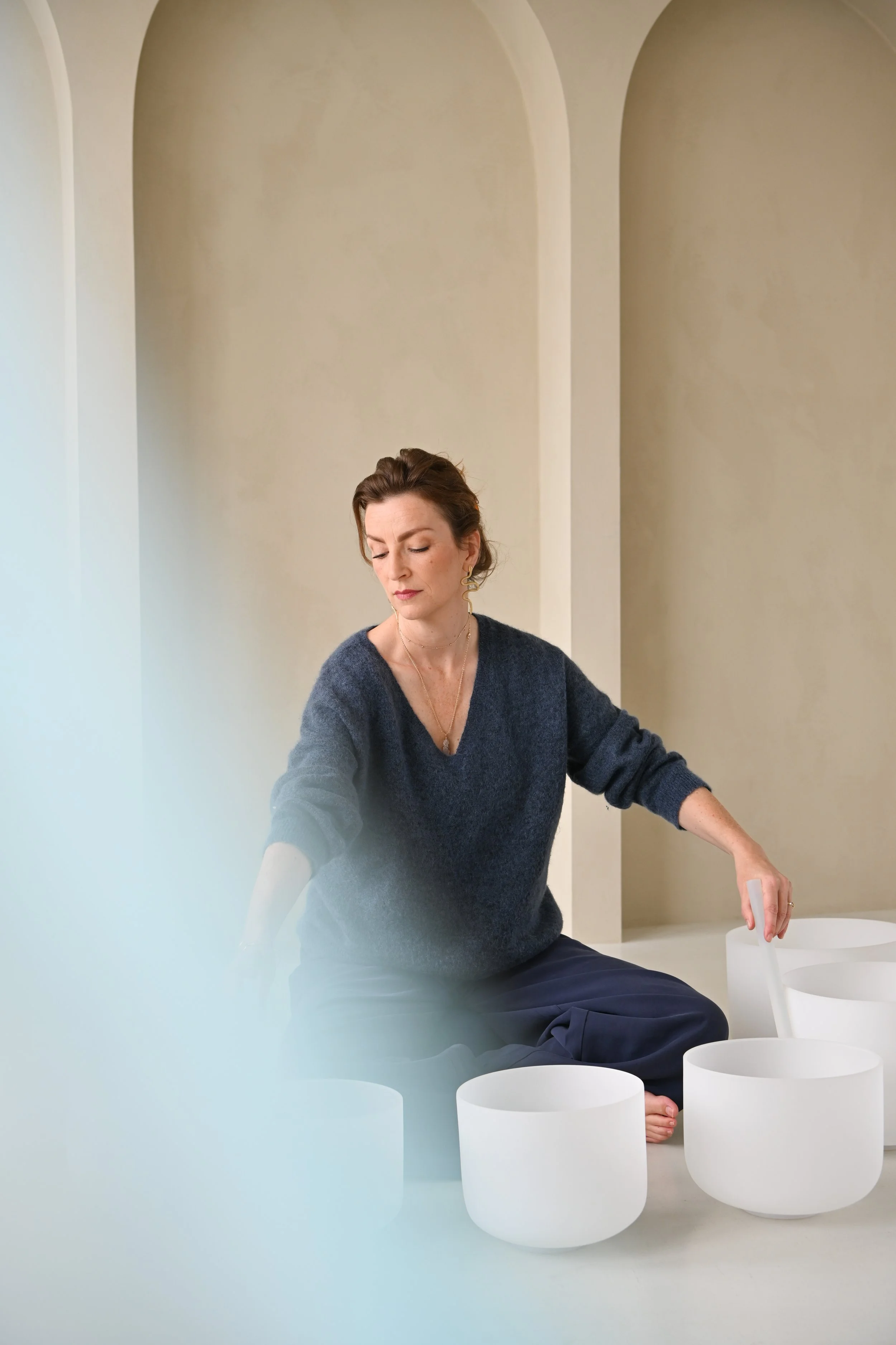 Woman sitting on the floor surrounded by white crystal singing bowls, with her eyes closed and hand reaching towards the bowls in a calm and meditative setting.