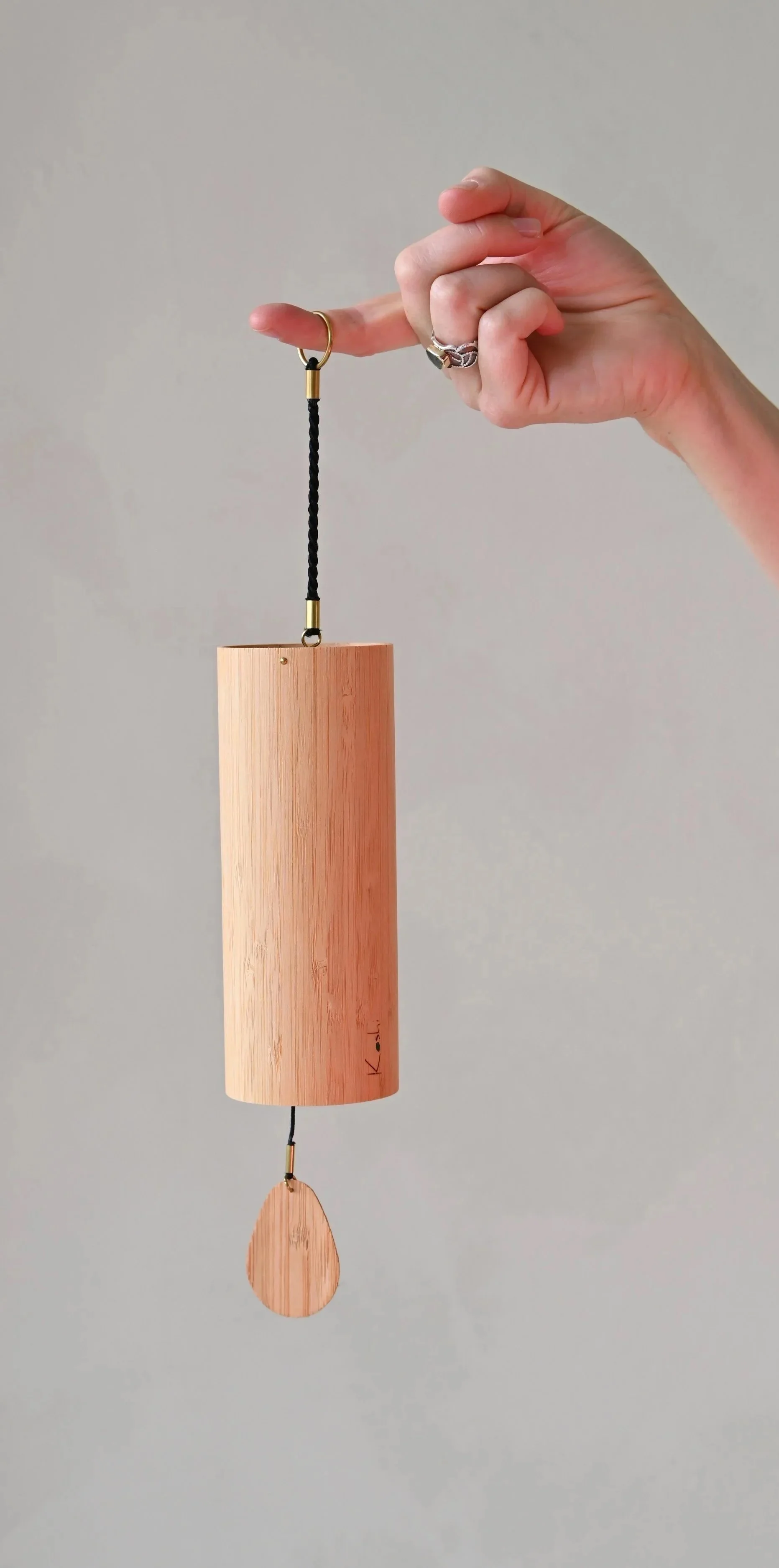 A person's hand holding a wooden wind chime with a black string, gold accents, and a teardrop-shaped wooden piece hanging at the bottom.