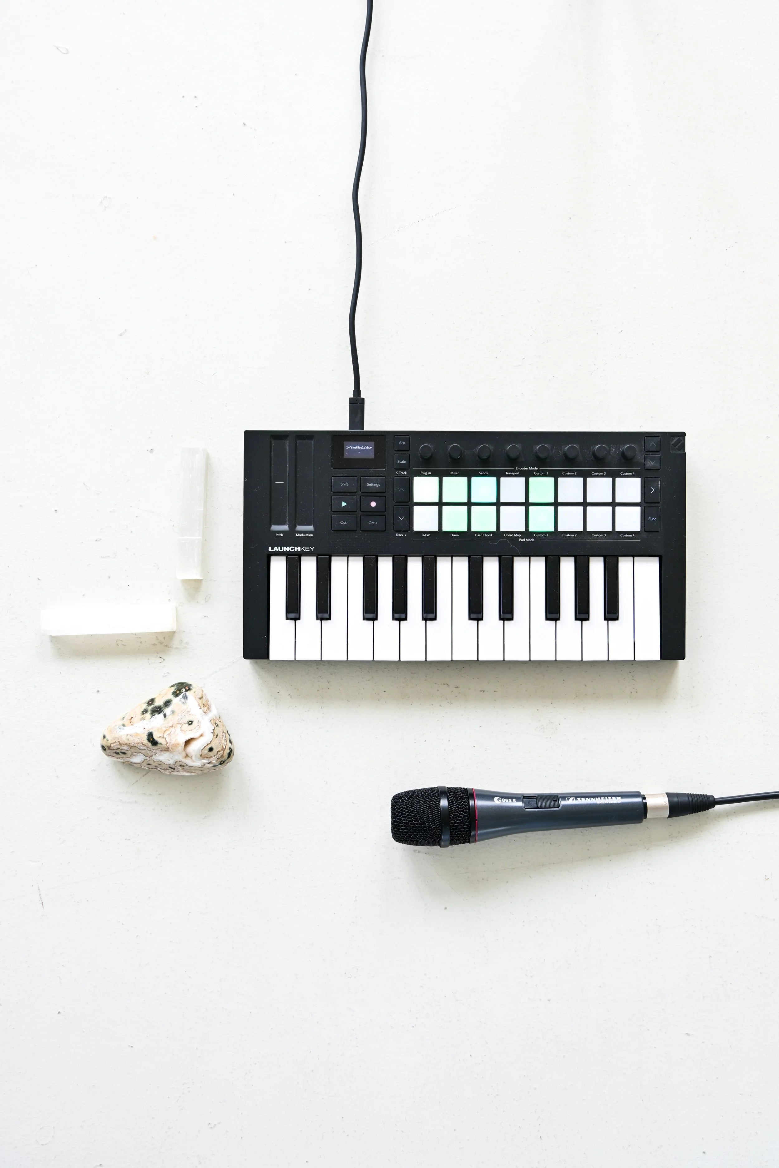 Music equipment including a small MIDI keyboard, a microphone, two white crystals, and a rock on a white surface.