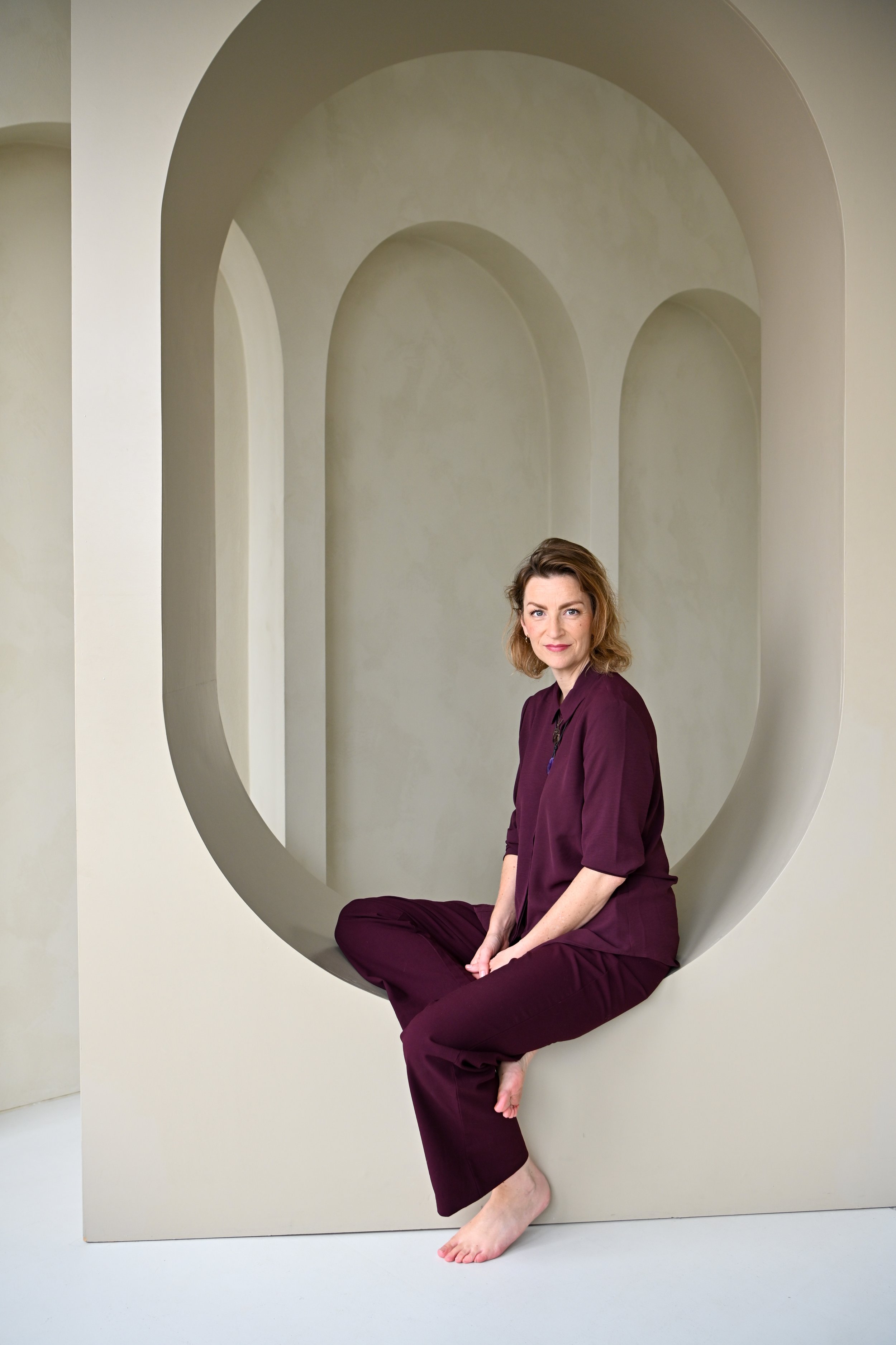 A woman in a burgundy outfit sitting cross-legged in a modern, minimalist architectural space with rounded arches and a neutral color palette.