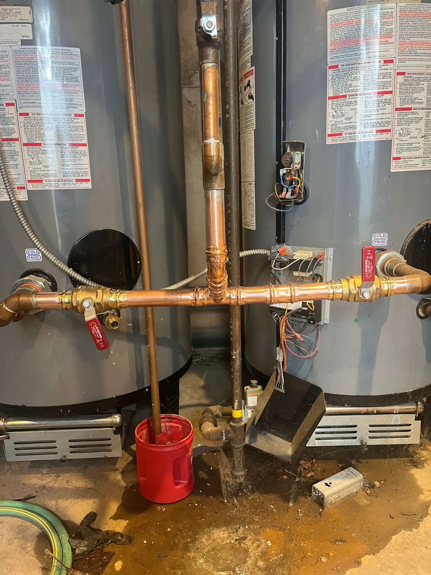A utility room with two large gray water heaters, copper pipes, a red bucket, and a small leak with water on the floor.