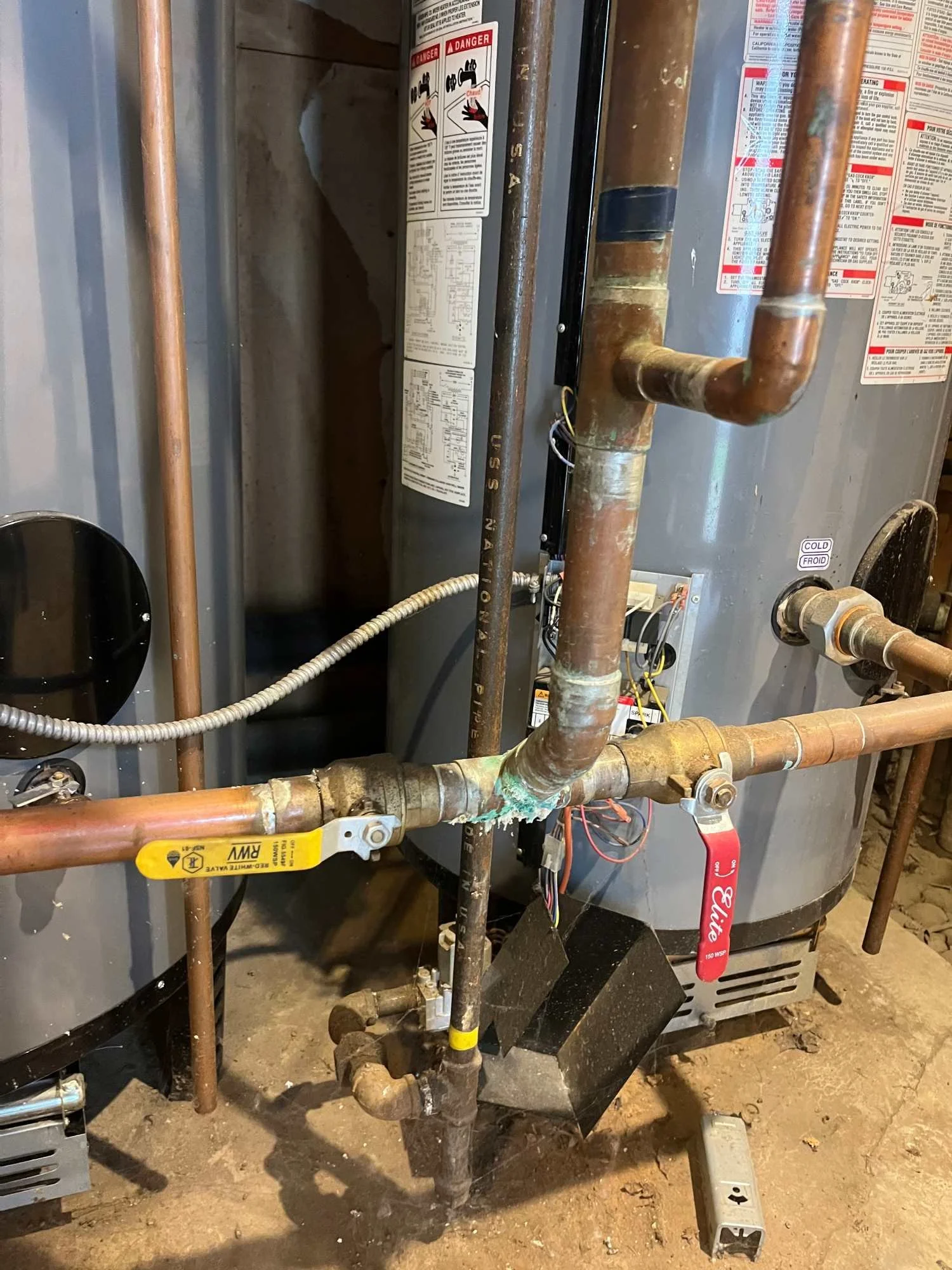 Close-up of a water heater with exposed copper pipes, some with corrosion and pipe fittings. Electrical wiring is visible near the base, and safety labels are attached on the heater.