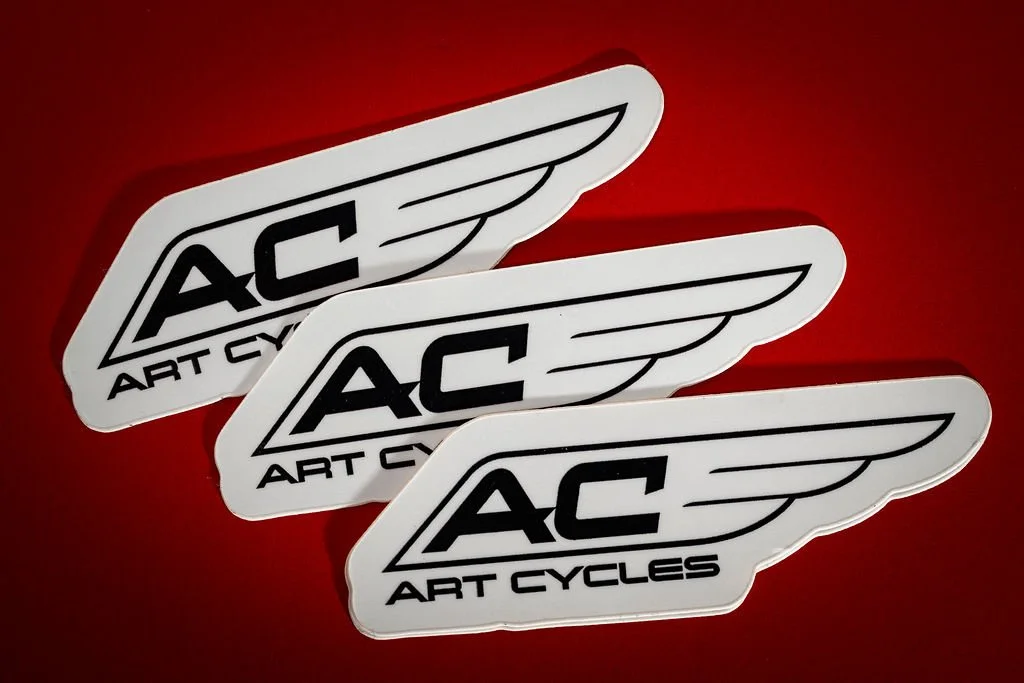 Three identical AC Art Cycles stickers with a winged logo on a red background.