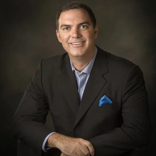 A man wearing a black suit with a blue shirt and pocket square, smiling for a portrait against a dark background.
