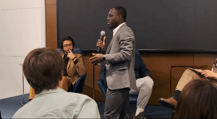 A man in a gray suit speaking into a microphone during a panel discussion in a conference room, with seated audience members and other panelists present.