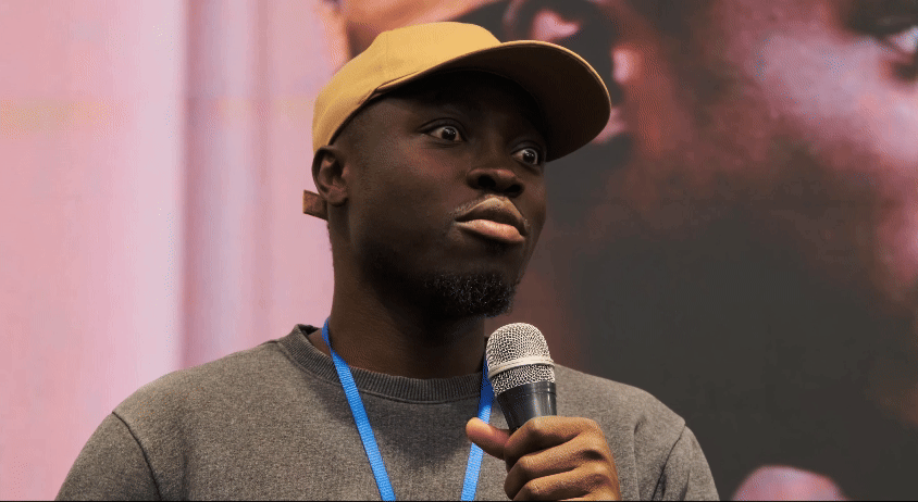 A young Black man wearing a beige cap and gray sweatshirt, holding a microphone, with a pink and black background.