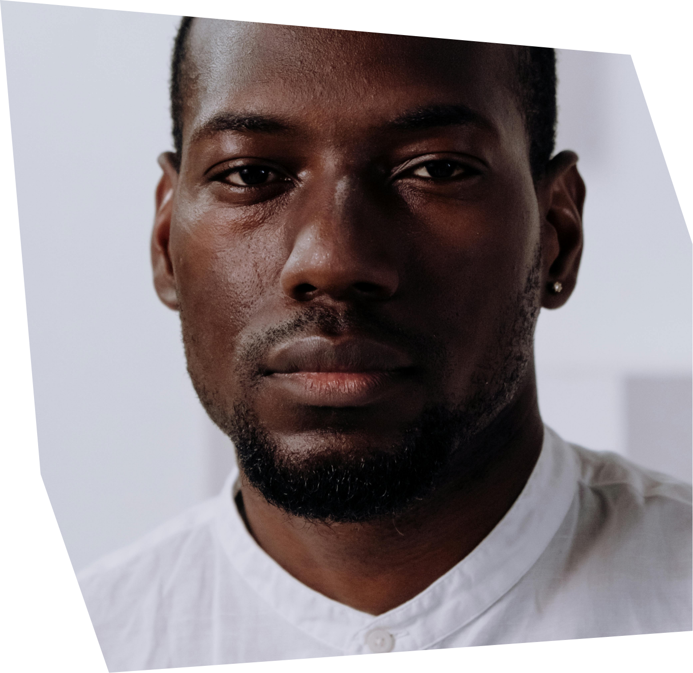 Close-up of a young Black man with short hair, a beard, and a white shirt, looking directly at the camera against a plain white background.