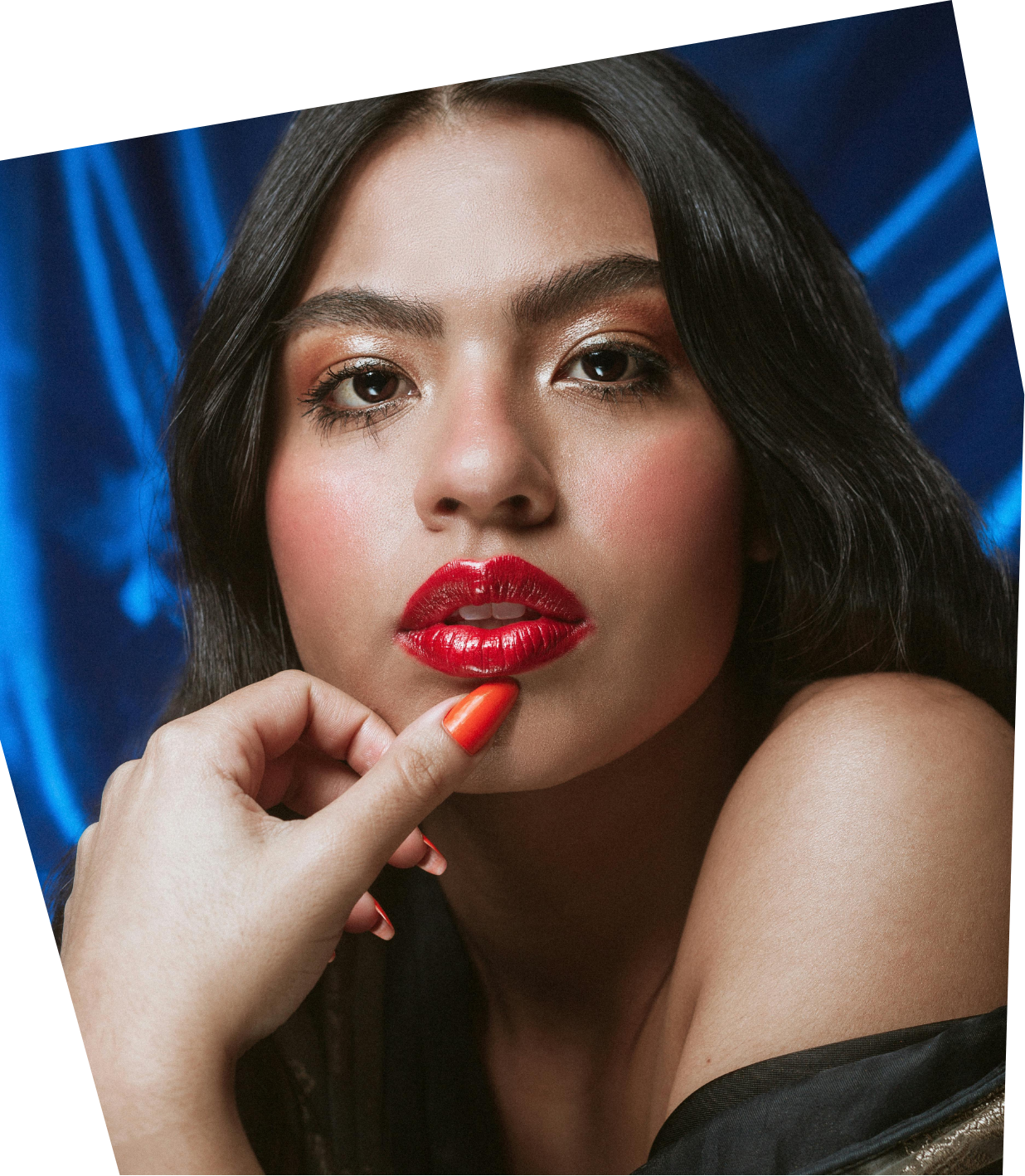 Close-up portrait of a woman with red lipstick, dark hair, and makeup, touching her chin with her hand against a dark blue background.