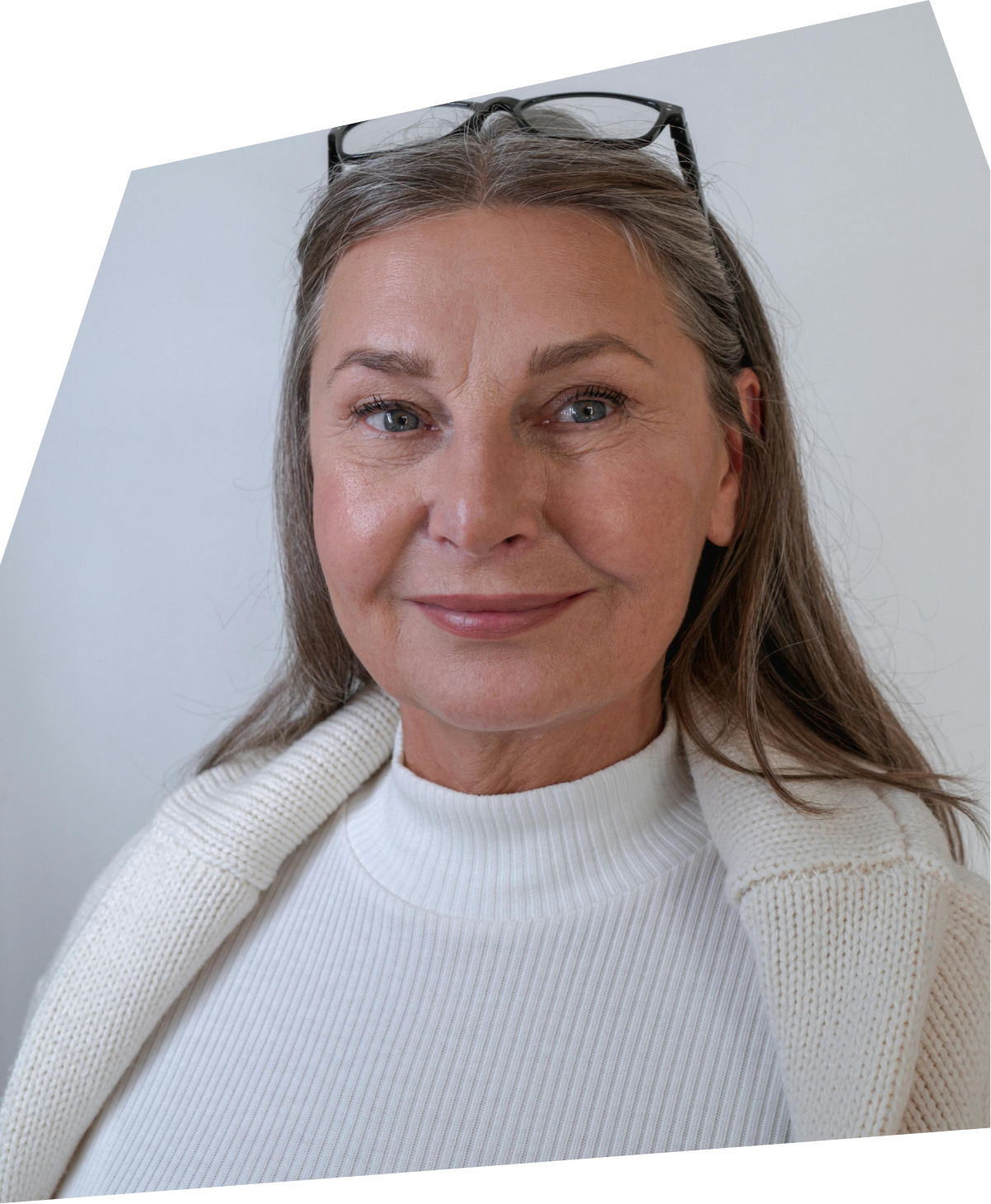 Close-up of a middle-aged woman with glasses on her head, smiling, wearing a white turtleneck sweater and a white knit scarf, against a plain light background.