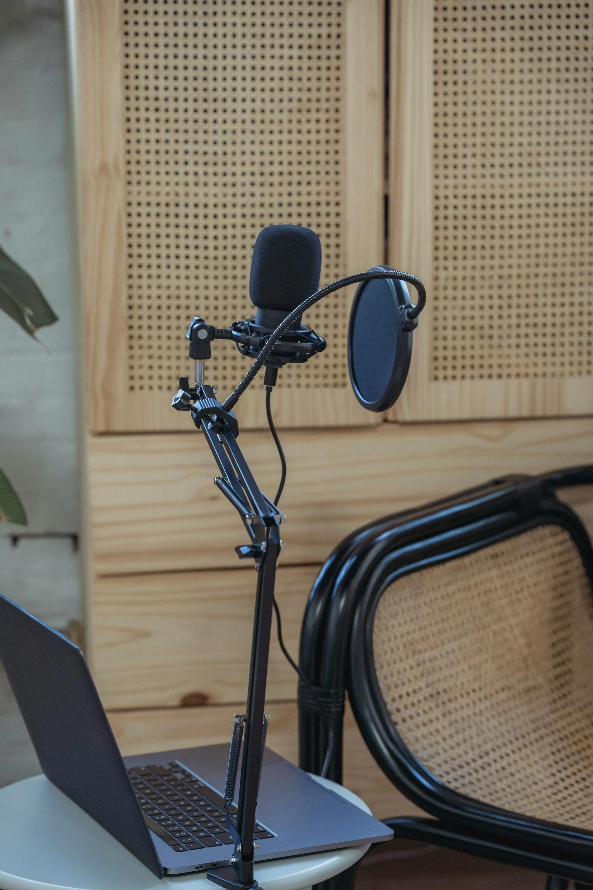 A microphone with a pop filter set up on a laptop in a recording studio or podcasting setup, with a woven wooden panel background and stacked chairs.