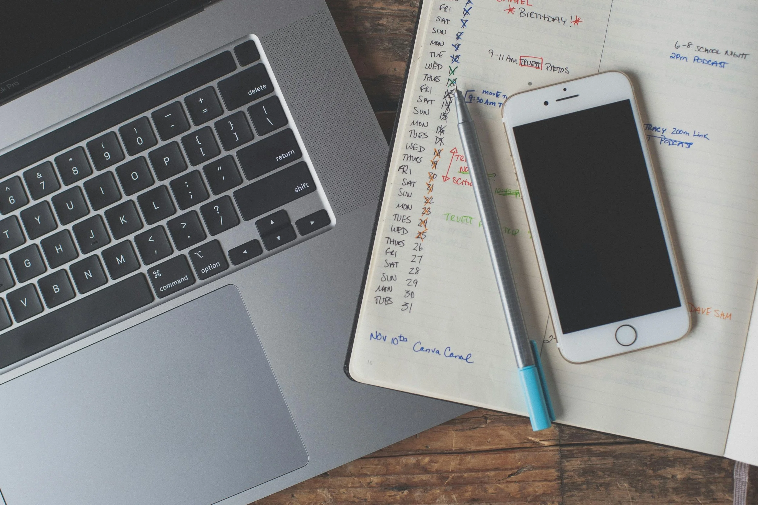 A laptop, a planner with handwritten notes and colored markings, a silver pen, and a white smartphone are arranged on a wooden surface.