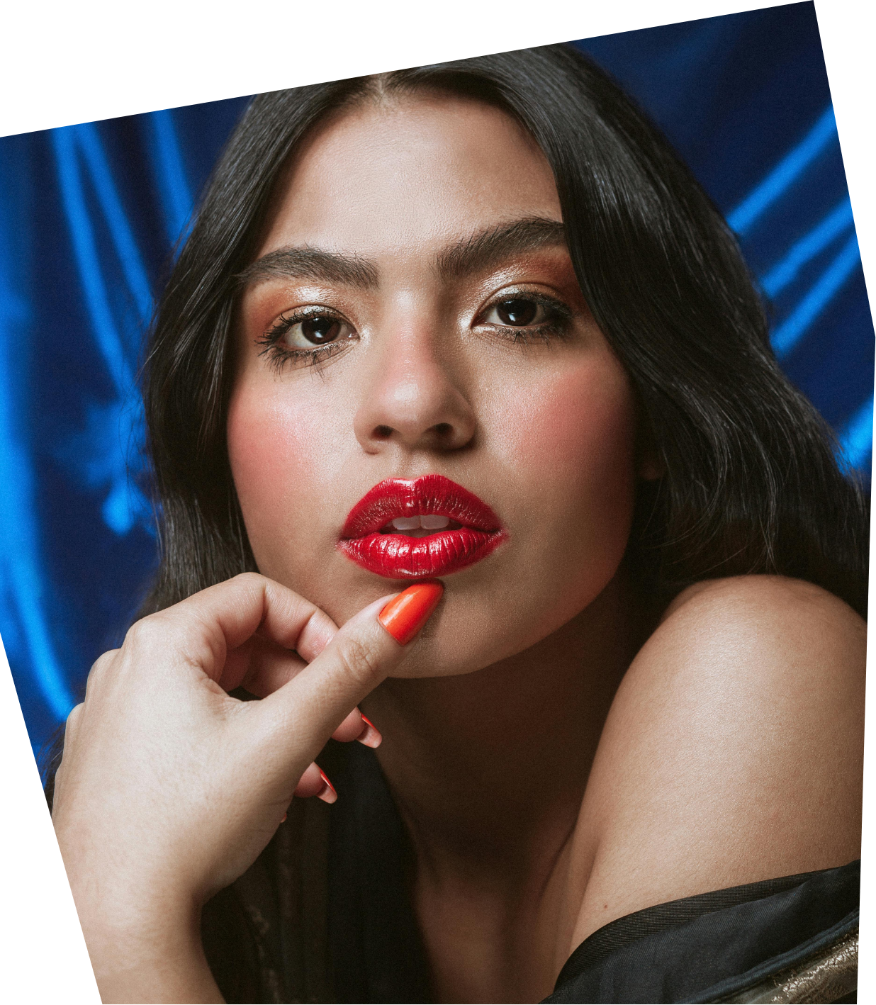 Close-up of a woman with red lipstick, dark hair, and makeup, resting her chin on her hand with orange-painted nails, against a blue satin backdrop.