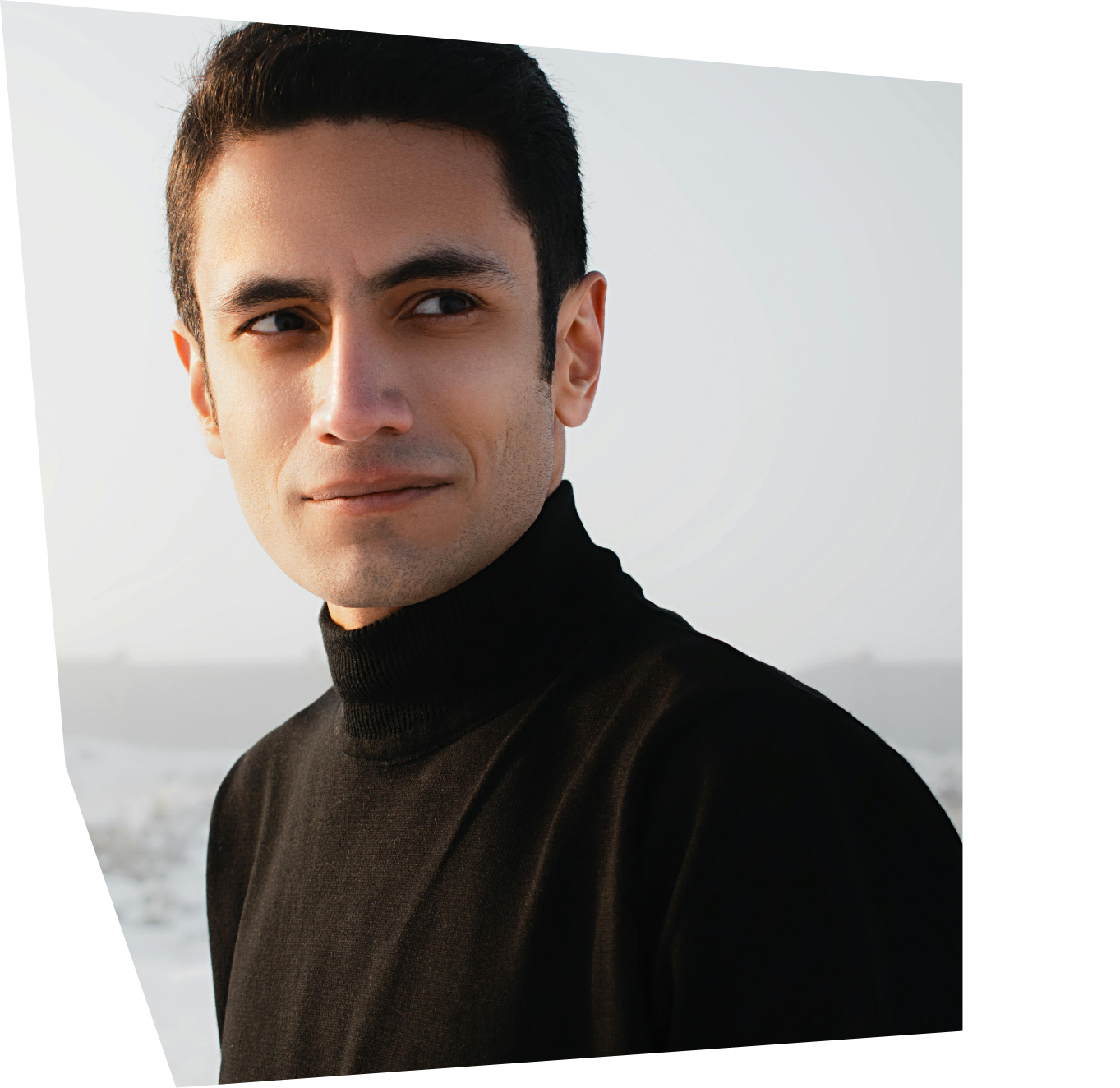 A young man with dark hair wearing a black turtleneck sweater outdoors with a beach and ocean in the background.