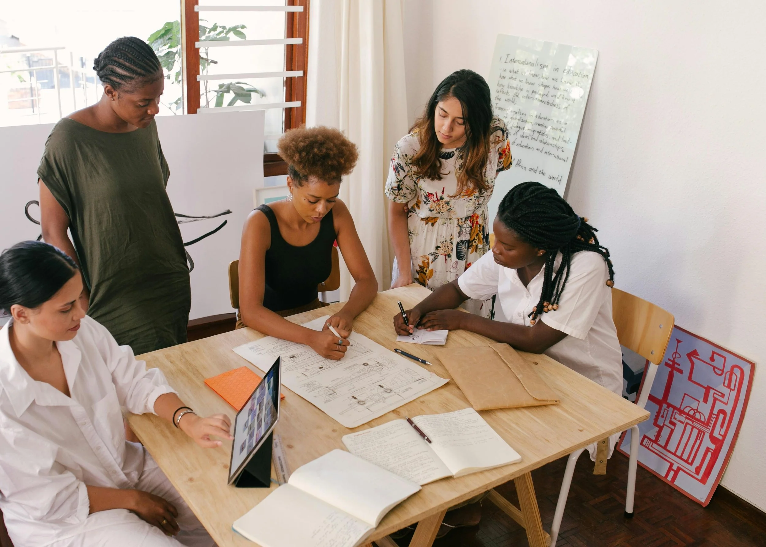 Group of five women collaborating around a wooden table, reviewing architectural blueprints and taking notes in a well-lit room with large windows and inspirational whiteboard.