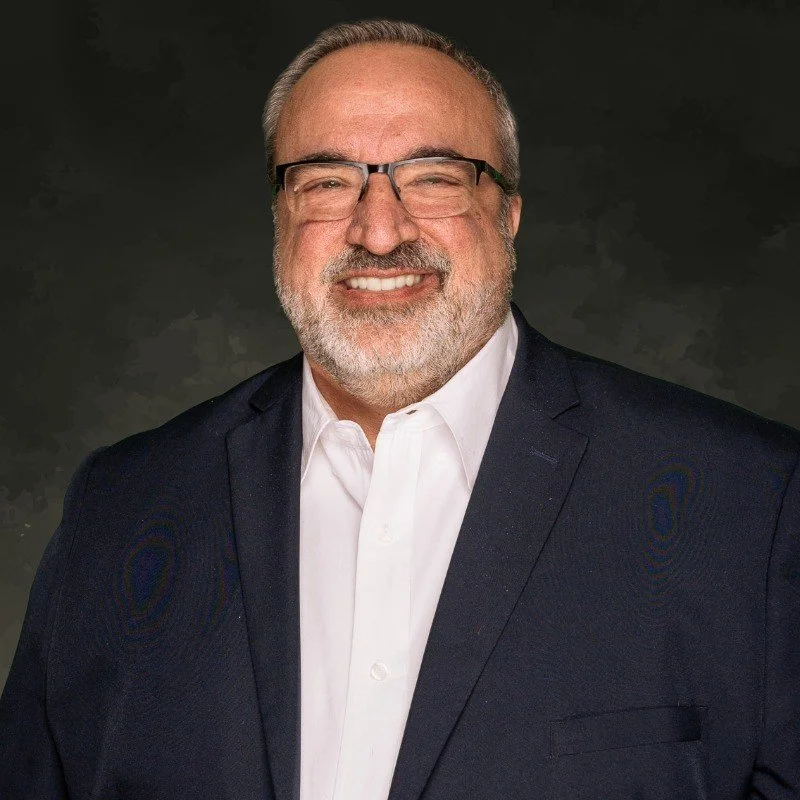 A smiling middle-aged man with gray hair, beard, and glasses wearing a white shirt and a dark suit against a dark background.