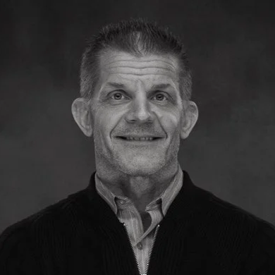 Black and white photo of a middle-aged man with short hair, wearing a dark zip-up sweater over a collared shirt, smiling slightly.