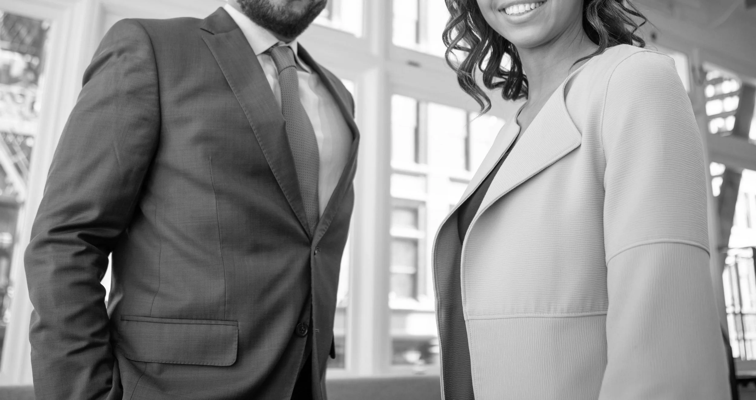 A man in a business suit and a woman in a blazer smiling at each other in an office setting.