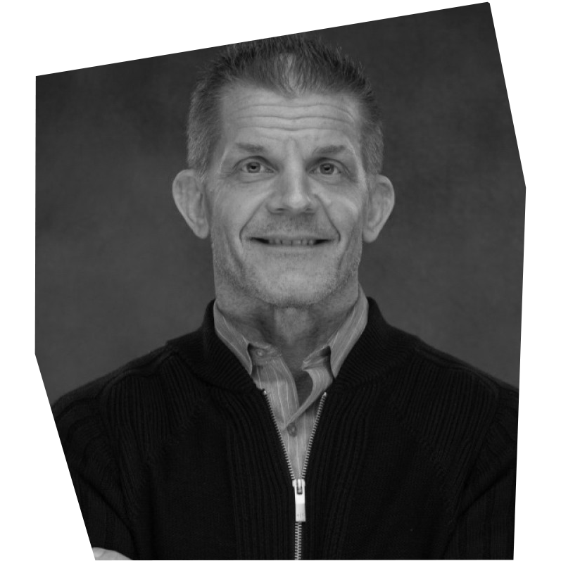 A black and white photo of a smiling middle-aged man with short hair, wearing a collared shirt and a zip-up sweater, against a plain background.