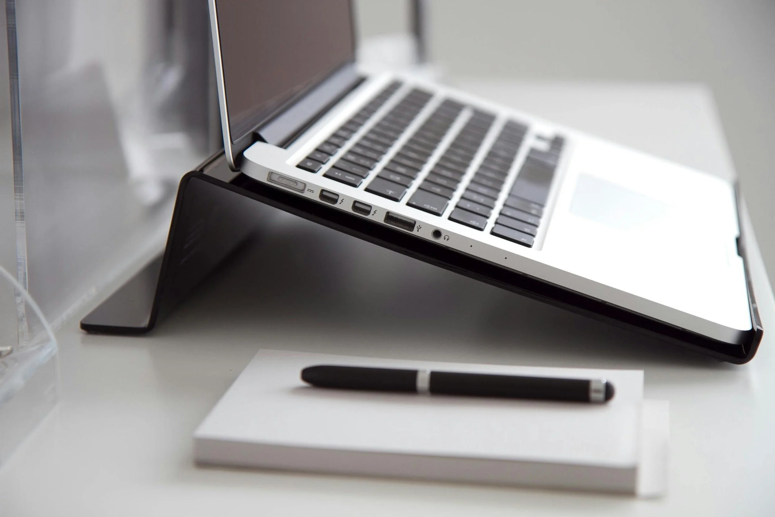 A close-up of a silver laptop with a black keyboard, placed on a white desk, propped up by a black stand, with a black pen resting on a white notebook nearby.