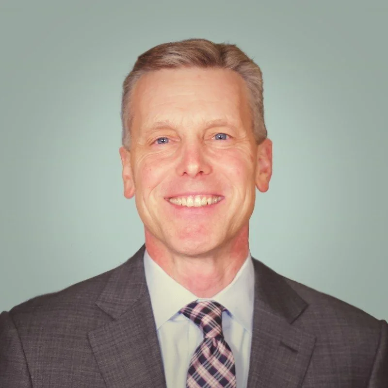 A professional headshot of a middle-aged man with short, light brown hair, blue eyes, wearing a gray suit, a white dress shirt, and a plaid tie, smiling against a light green background.