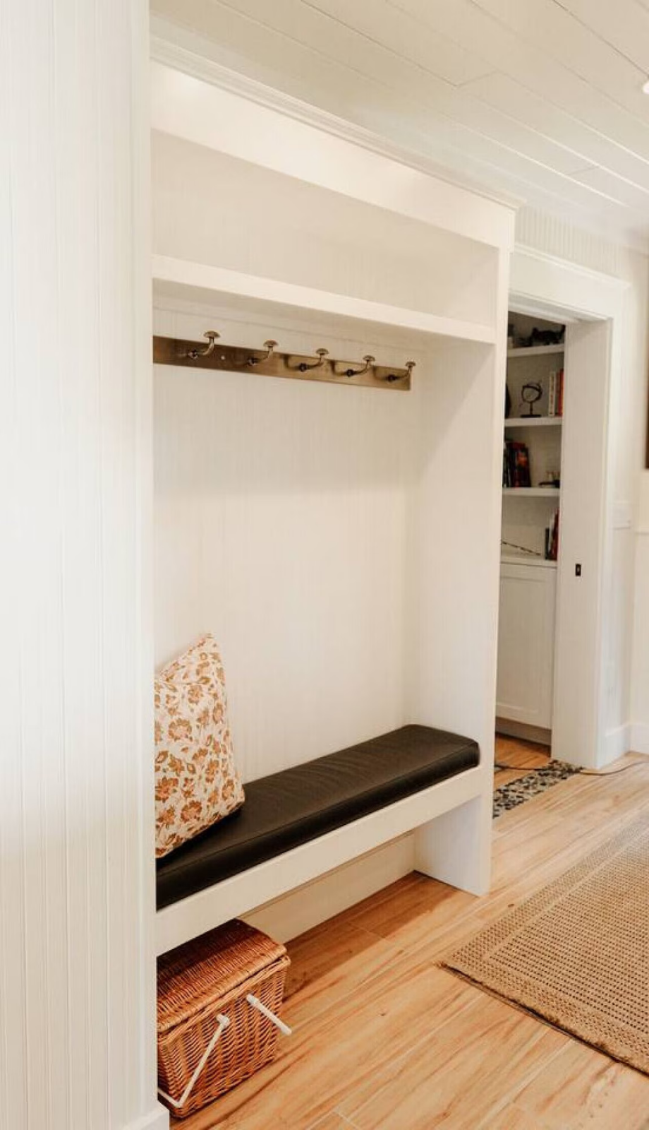 Empty built-in entryway nook with a black cushion bench, a floral pillow, a wicker basket below, and a coat rack on the wall, with a bookshelf in the background.
