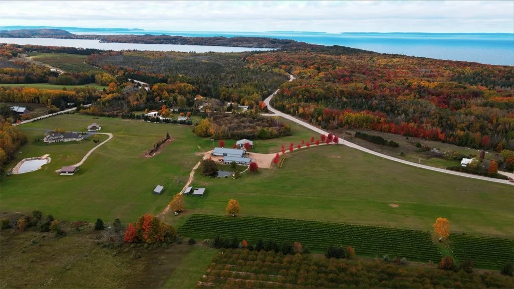 Aerial view of a rural landscape with fields, trees, a winding road, farm buildings, and a large body of water in the background, showcasing fall foliage.