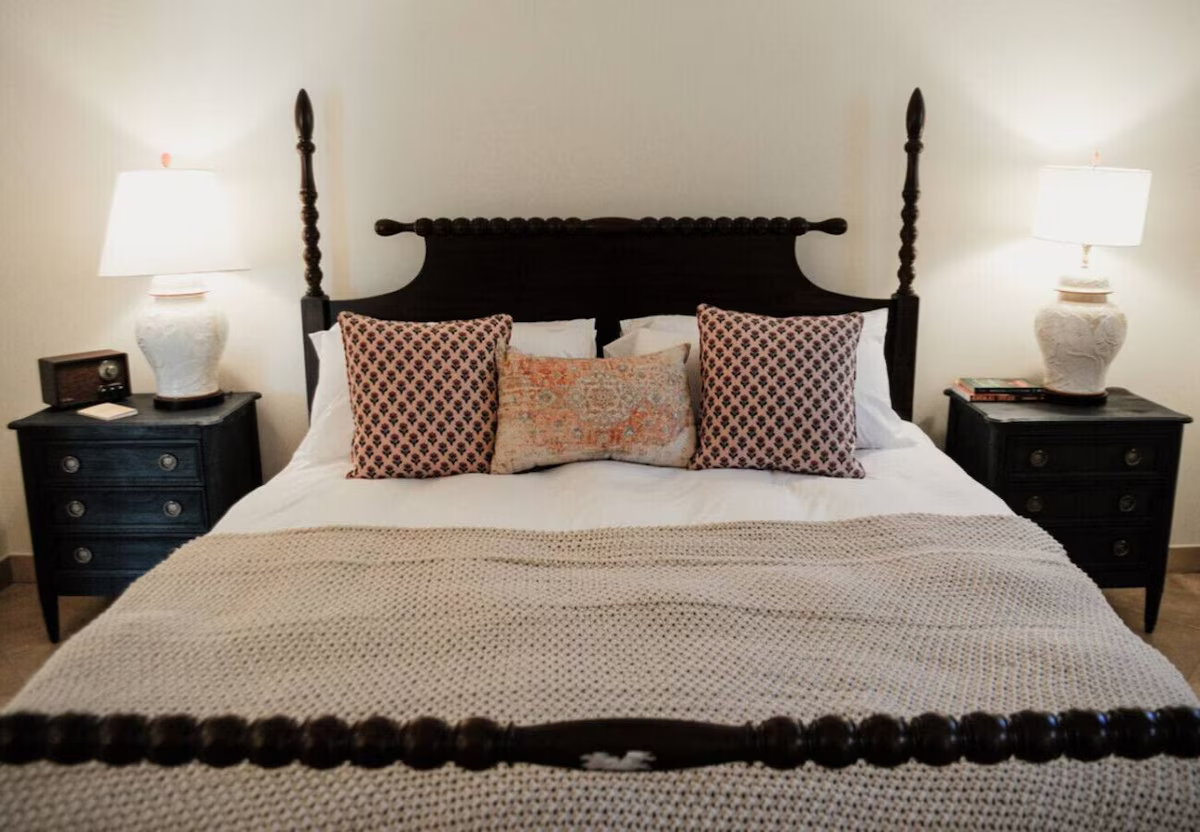 A neatly made bed with a dark wooden four-poster headboard, adorned with decorative pillows and a beige knitted blanket. Flanking the bed are two black nightstands, each with a large white ceramic lamp and some books. The wall behind is plain and lig
