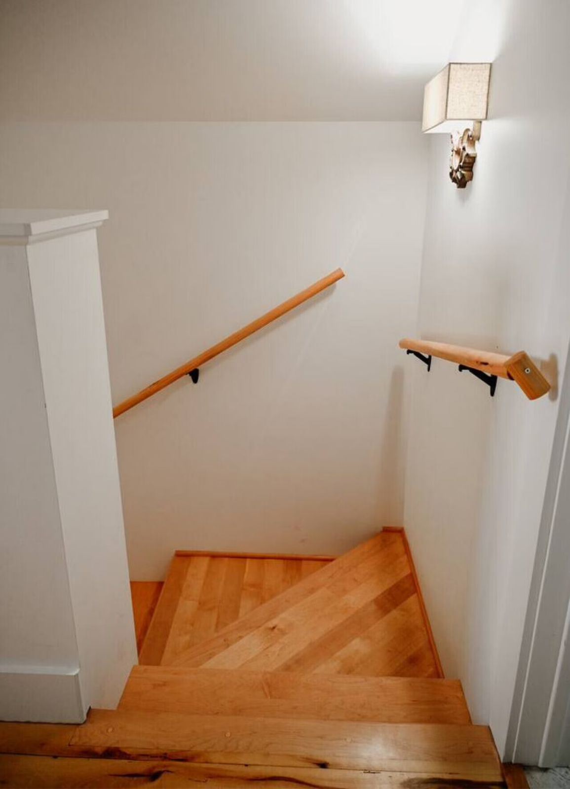 Wooden staircase with handrails on both sides, white walls, and a wall-mounted light fixture.