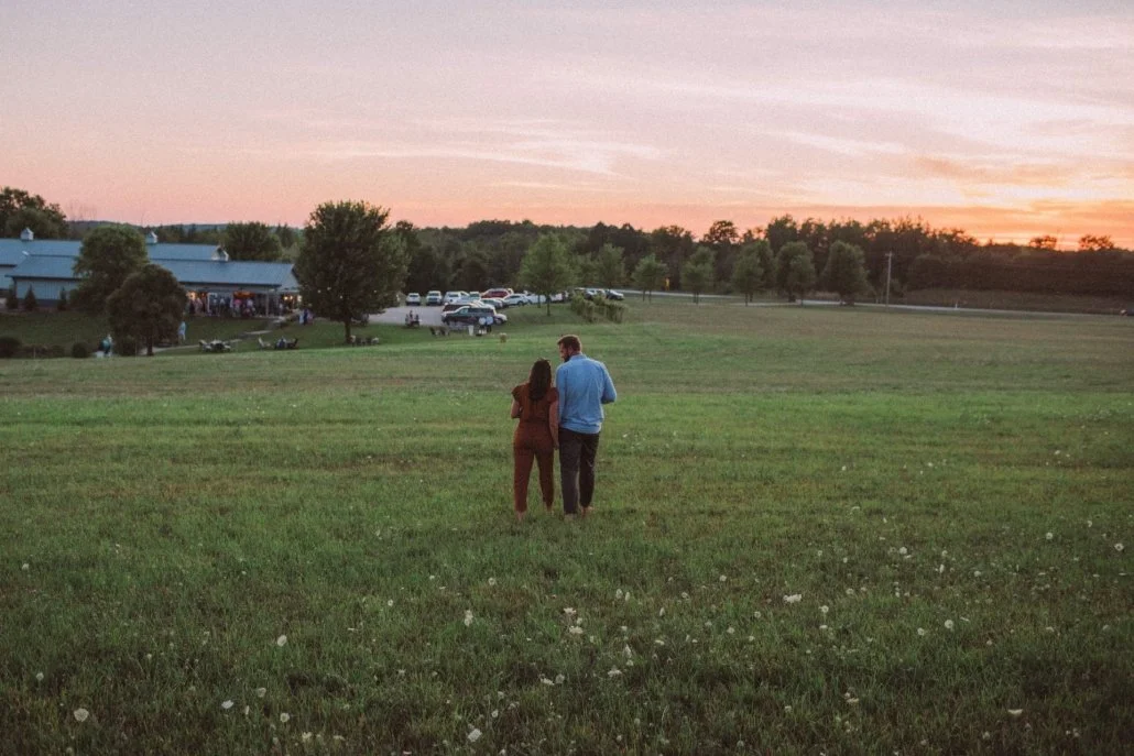 A couple walking together on a grassy field at sunset, with a building, parked cars, and trees in the background.
