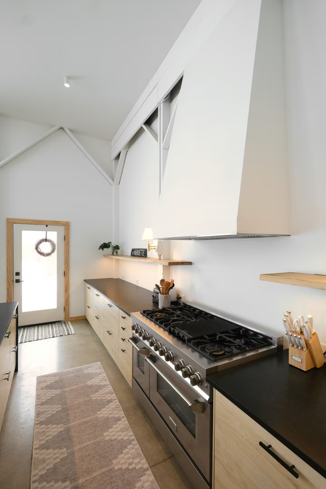 Modern kitchen with black countertops, stainless steel oven, white cabinets, and wooden accents. There is a door with a wreath, a small shelf with a lamp, and a countertop with kitchen utensils.