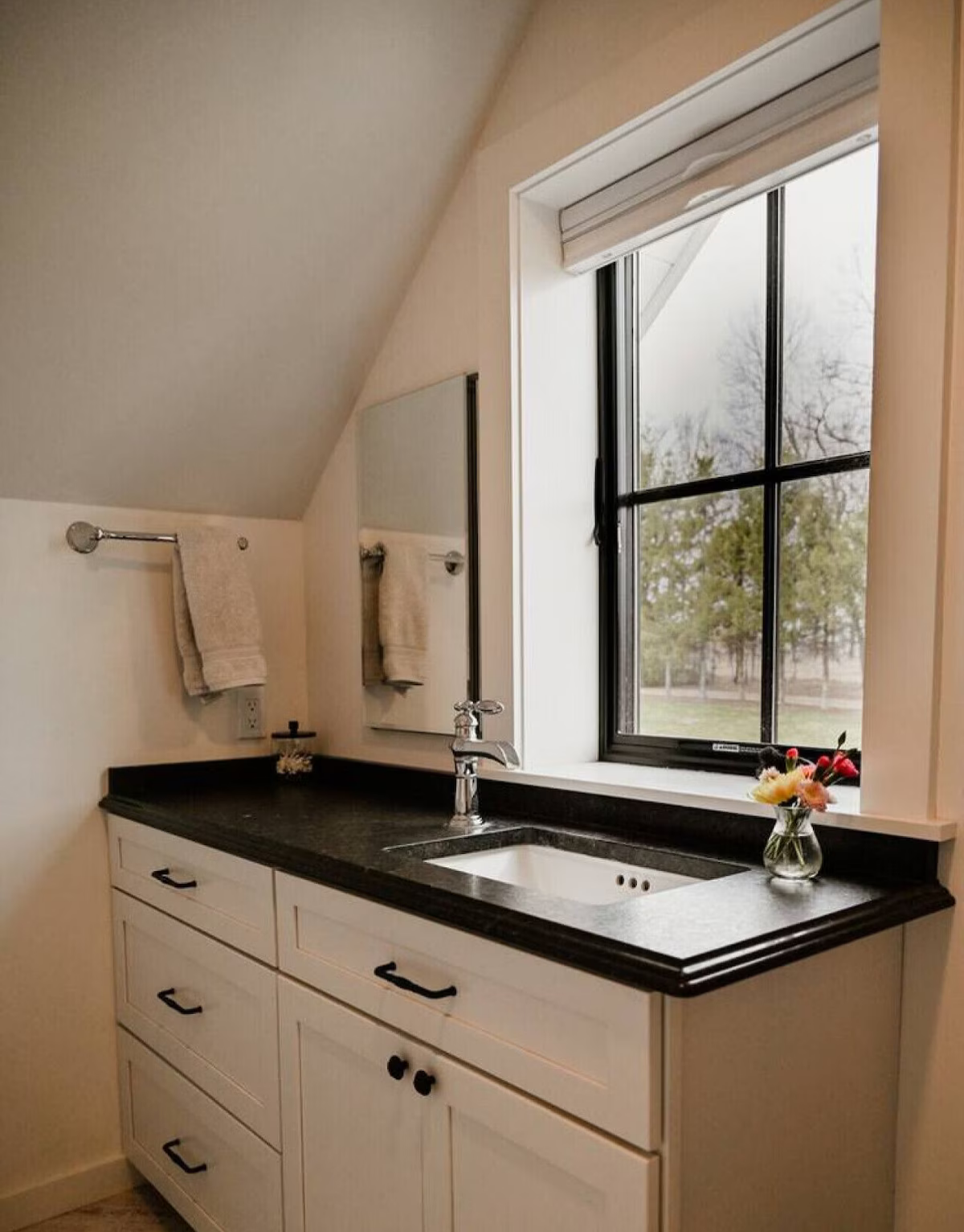 Bathroom with black countertop, white cabinet, window with view of trees, towel rack with towels, small vase with flowers