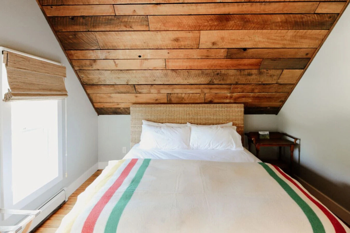 Bedroom with a bed, wooden headboard, and slanted wooden ceiling, next to a window with a beige Roman shade.