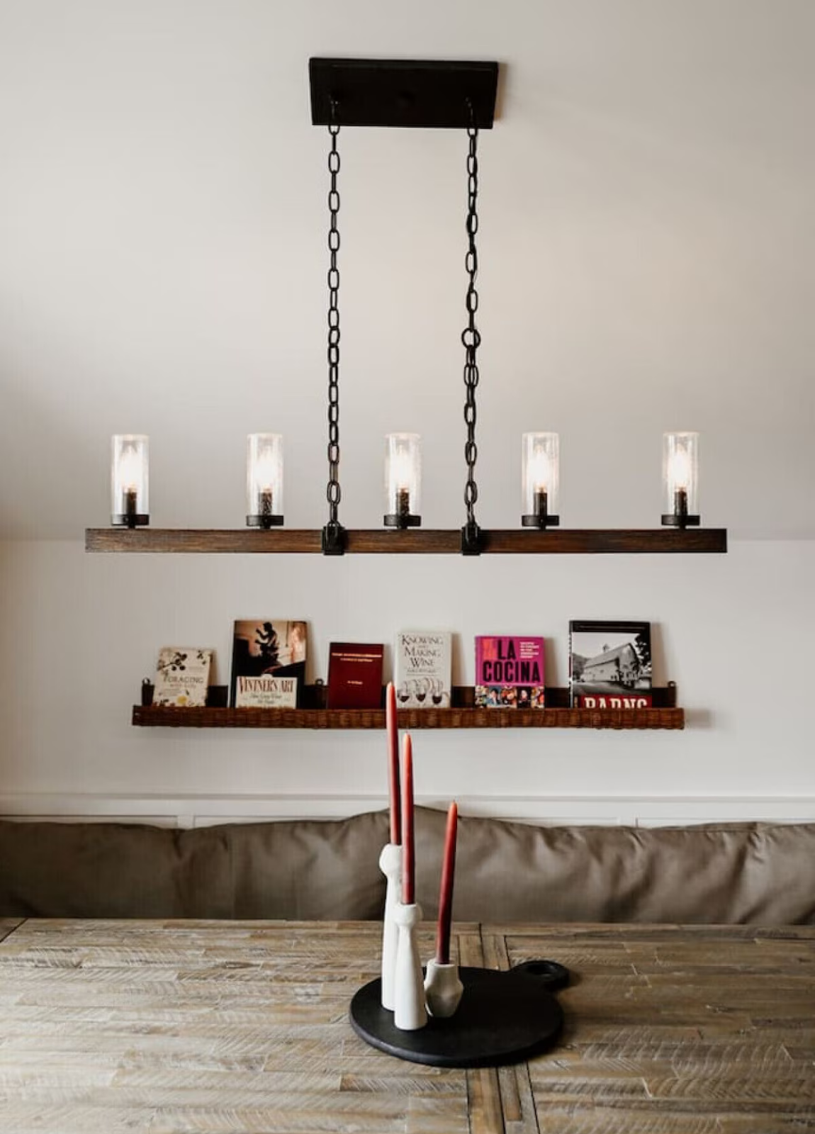 A dining room with a wooden table, candle holders with red candles, a white cloth on the candles, and a black tray. The wall has two wooden shelves with books and decorative items, and a modern black chain chandelier with exposed light bulbs hanging 