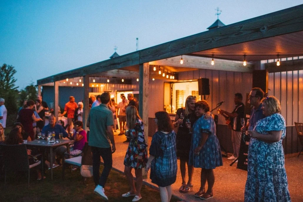 People gathering and socializing at an outdoor evening event with string lights, a DJ or musician performing, and a wooden pavilion.