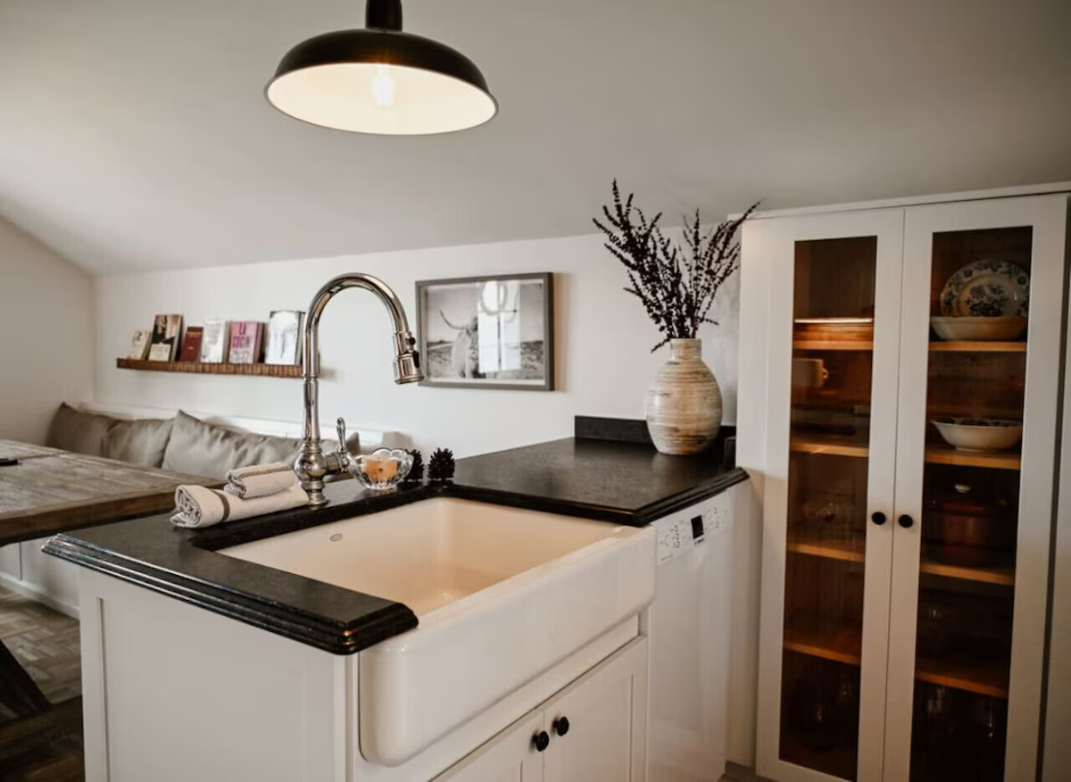Kitchen with white cabinets, black countertop, and farmhouse sink, with a vase of dried flowers and built-in bookshelf in the background.