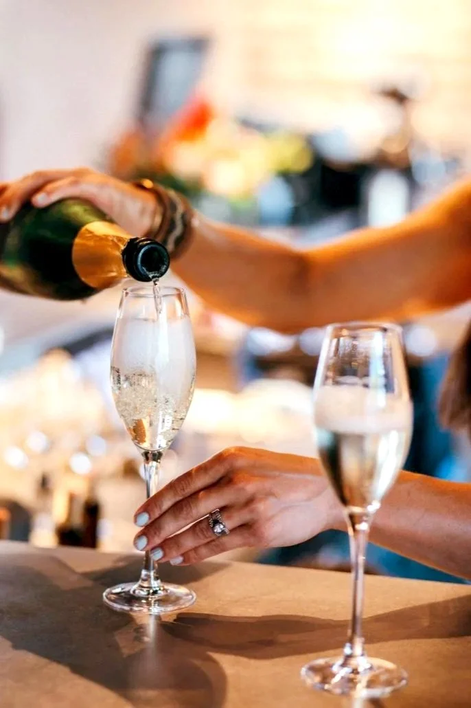 Person pouring champagne into a tall flute glass, with another filled glass nearby on a kitchen counter.