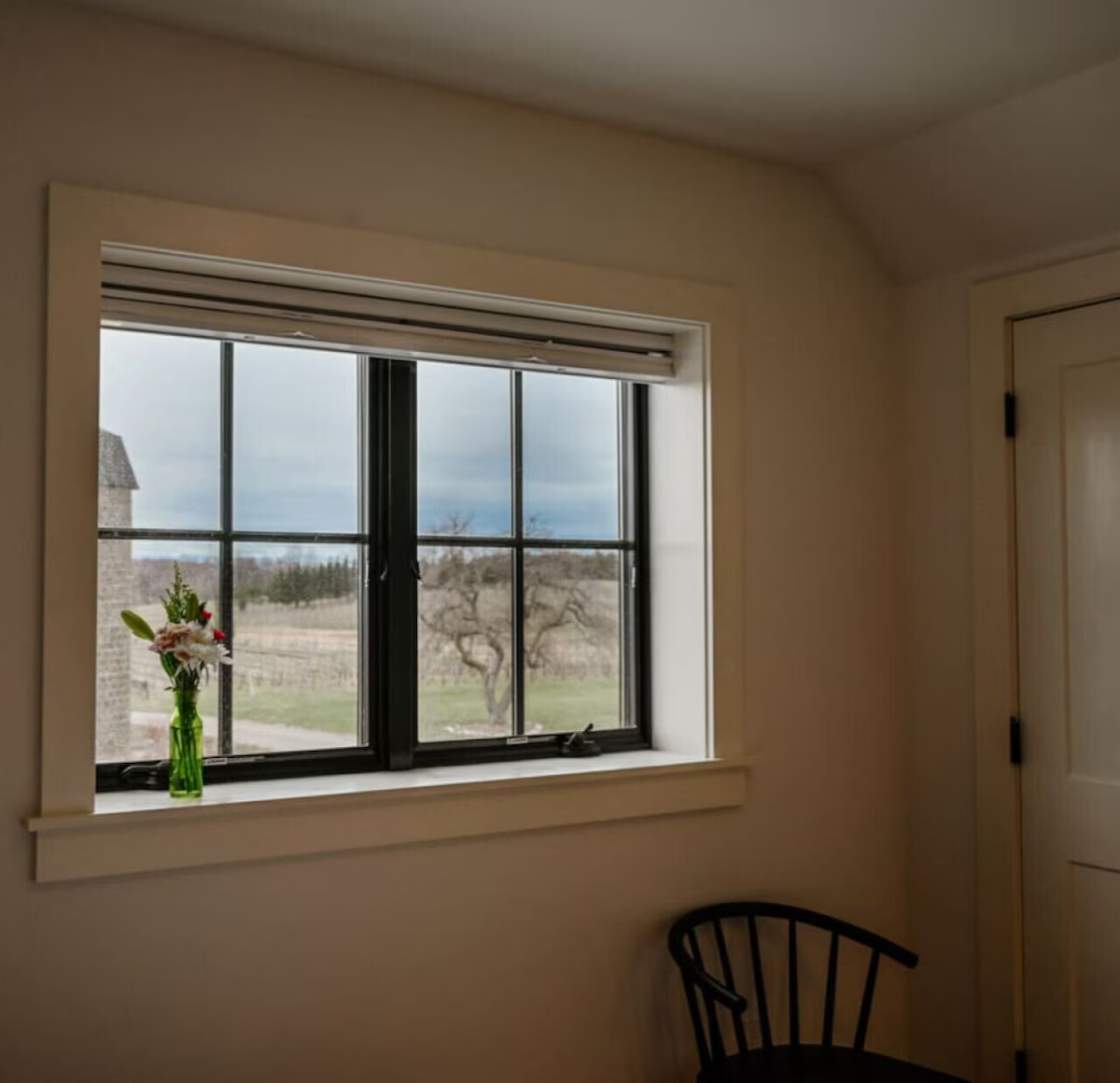 Interior of a room with a window showing an outdoor landscape with trees and a cloudy sky, a green vase with flowers on the windowsill, and a black chair partially visible in the lower right corner.