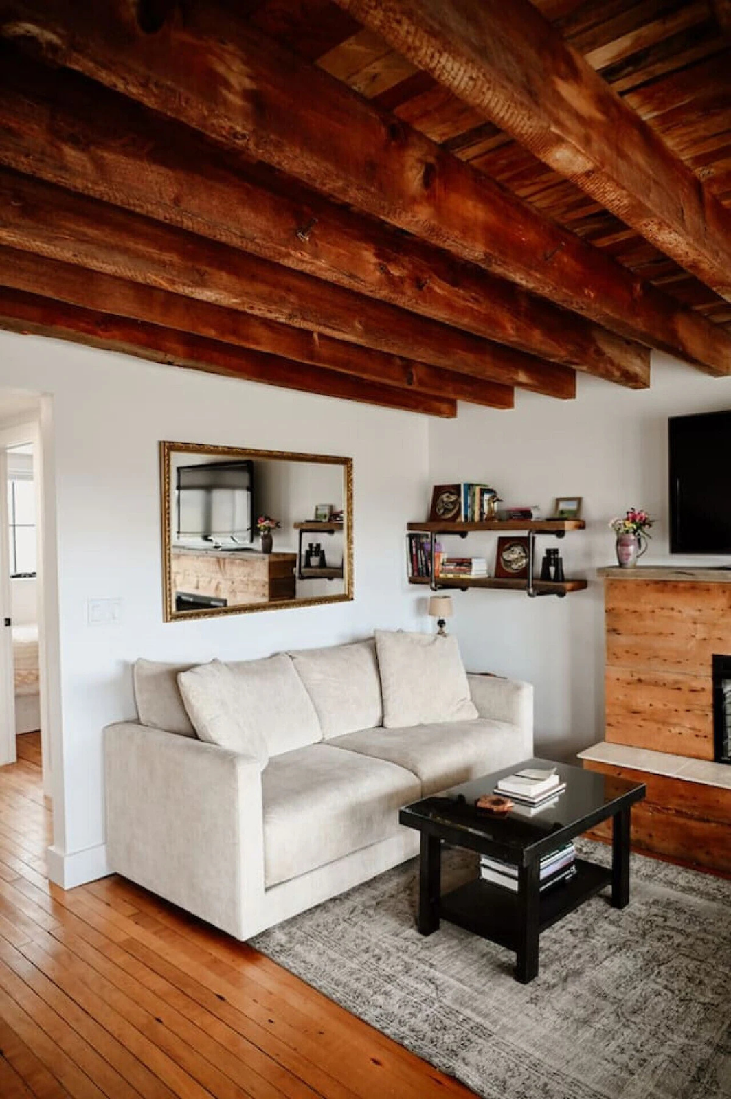Living room with white sofa, wooden ceiling beams, wall mirror, TV, wall shelves with books and decorative items, fireplace, and area rug.